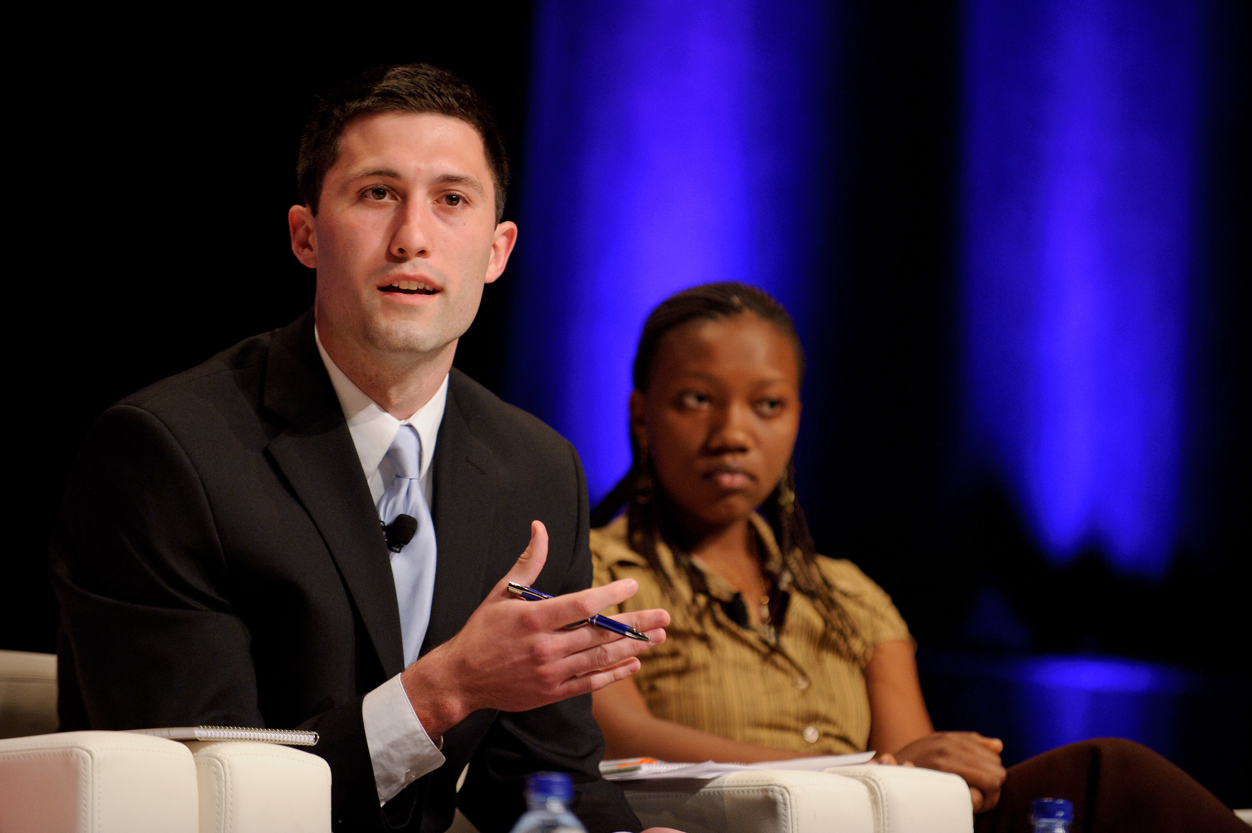 A man in a suit speaks while seated on a panel next to a woman, with blue lights in the background.