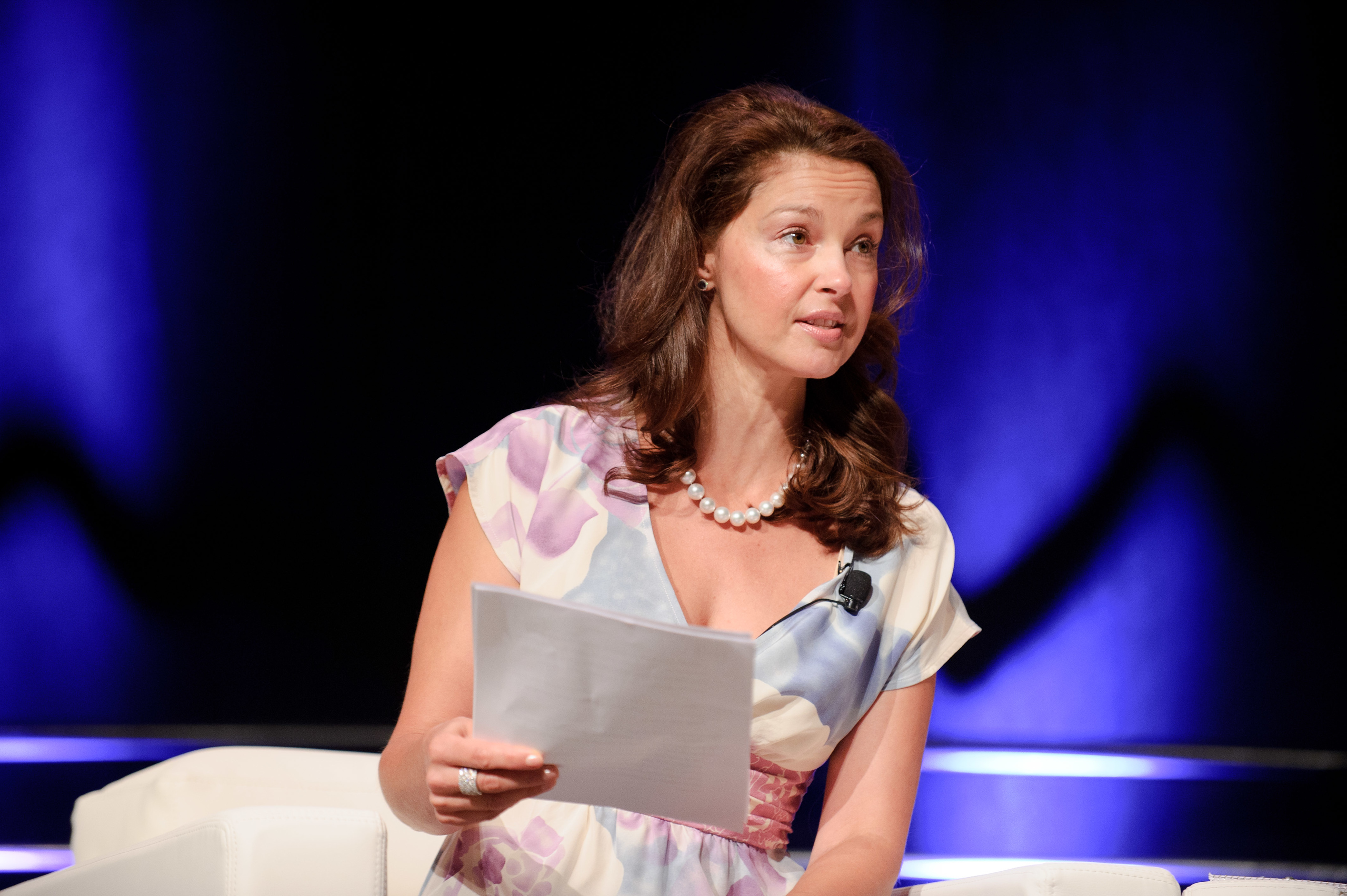 A woman in a floral dress sits on a white chair, holding papers and speaking, with a blue and purple-lit background.