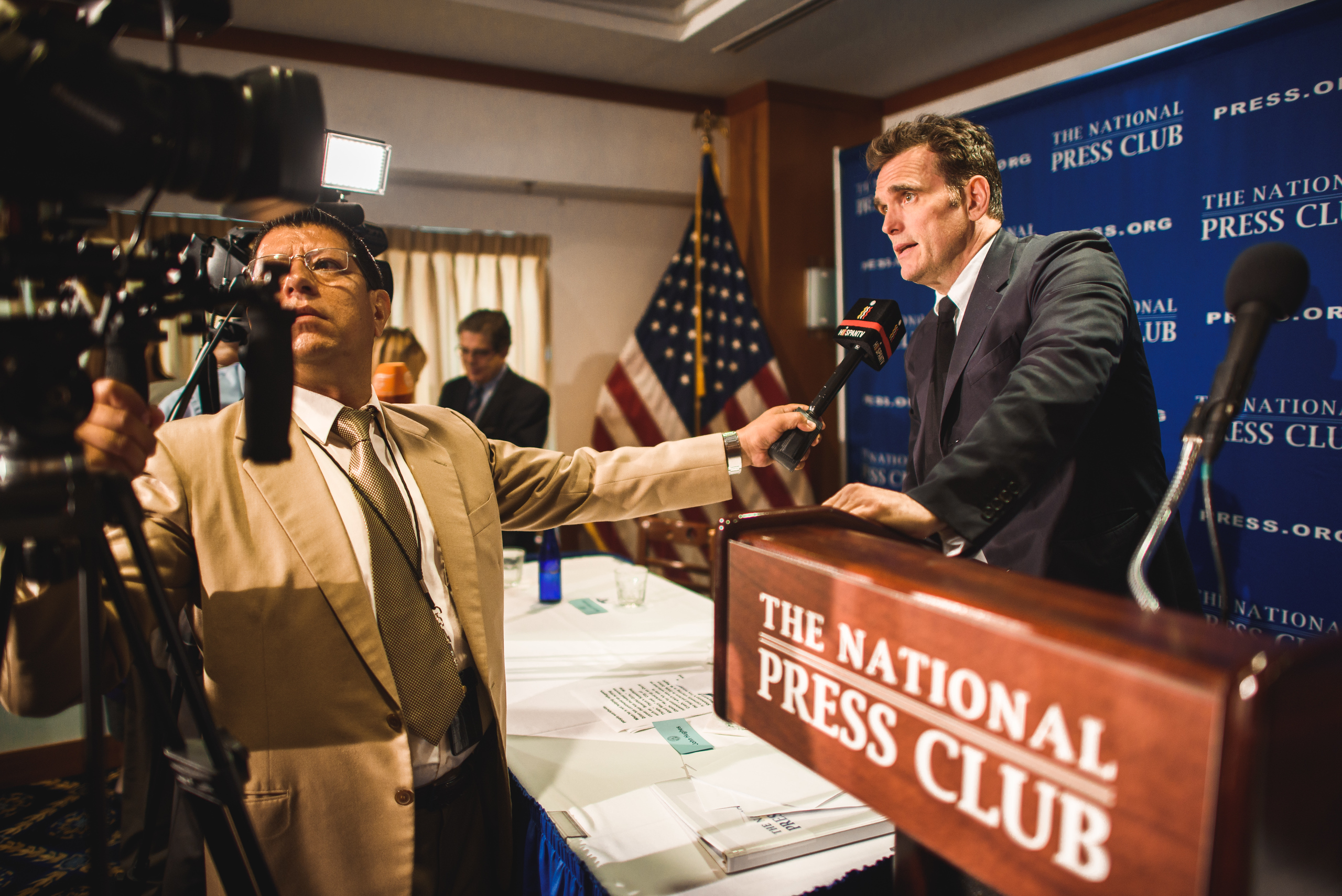 A man speaks at a podium labeled "The National Press Club" while reporters with microphones and cameras cover the event. An American flag is visible in the background.