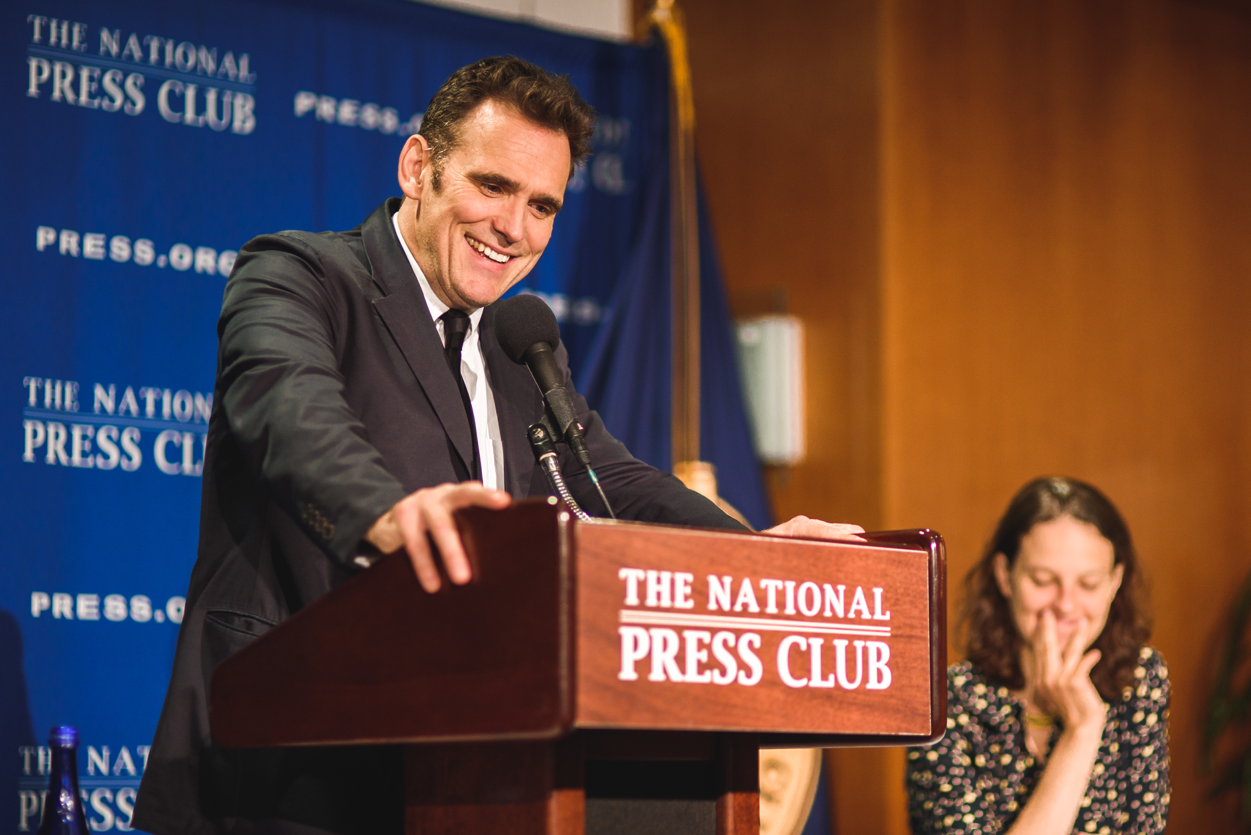 A man in a suit speaks at the National Press Club podium, smiling, while a woman seated nearby laughs with her hand over her mouth.