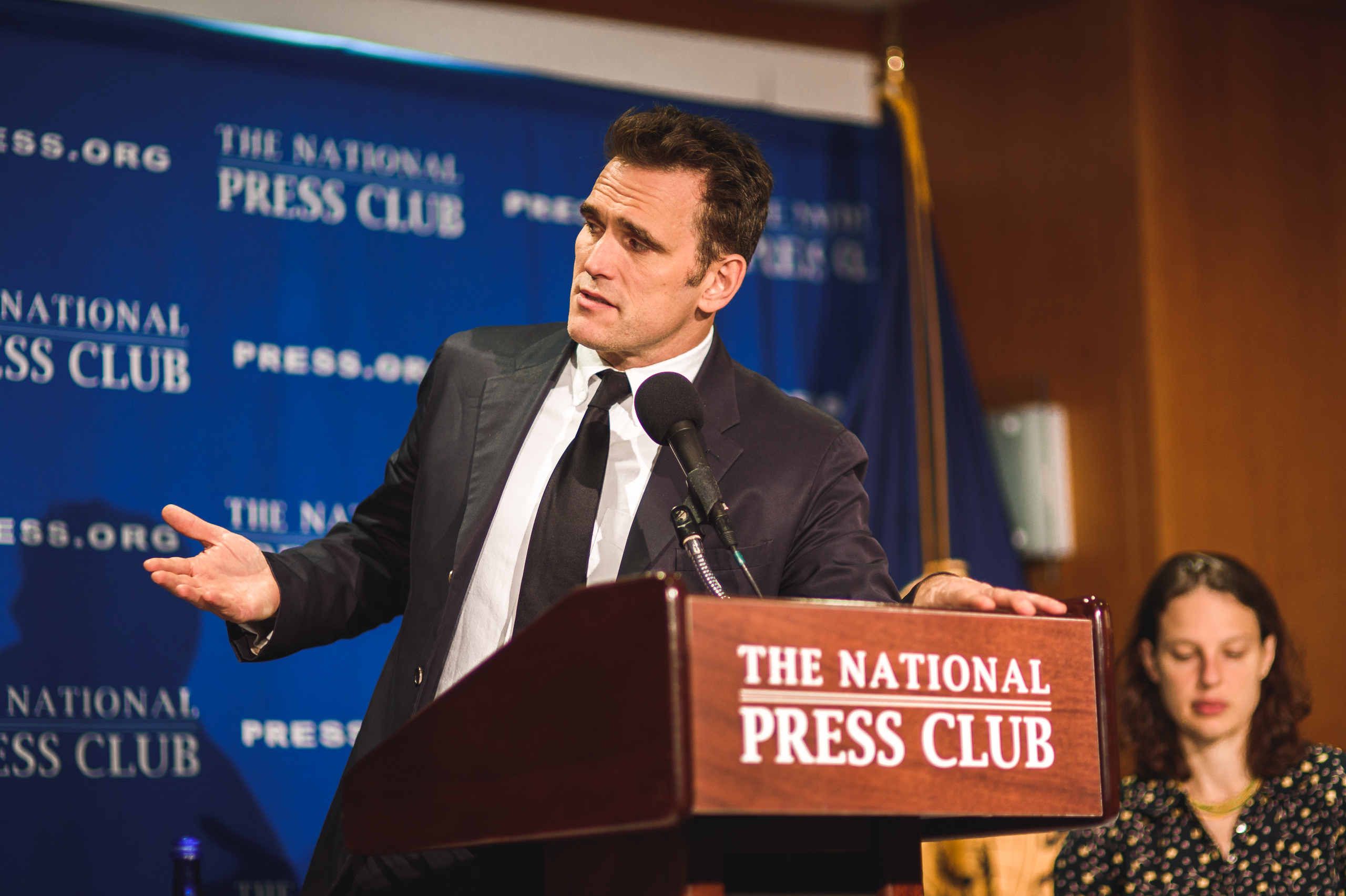 A man in a suit speaks at a podium labeled "The National Press Club" with a woman seated behind him and a blue press club backdrop.
