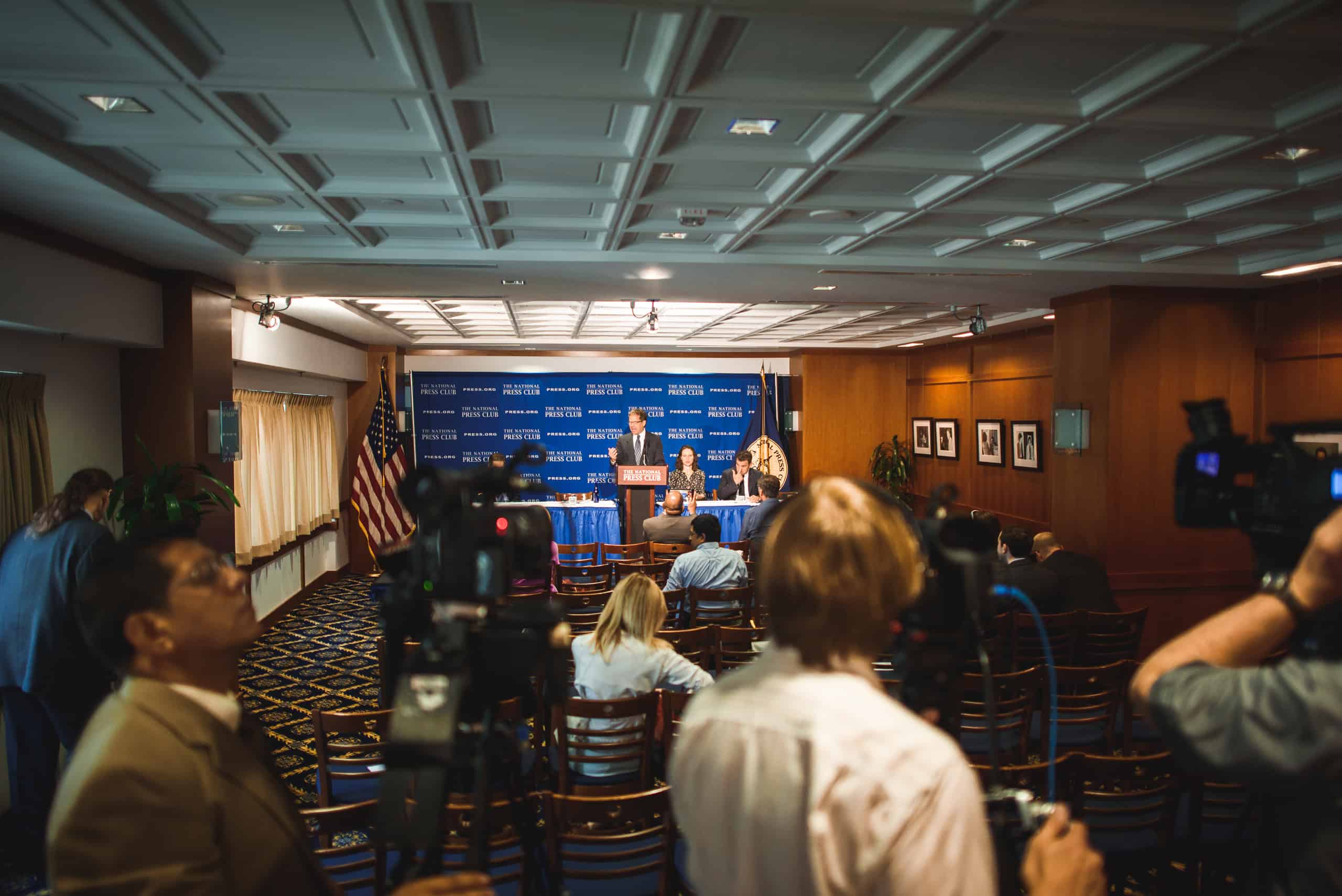 A press conference with speakers seated at a table onstage, journalists and cameras in the foreground, and an American flag in the background.