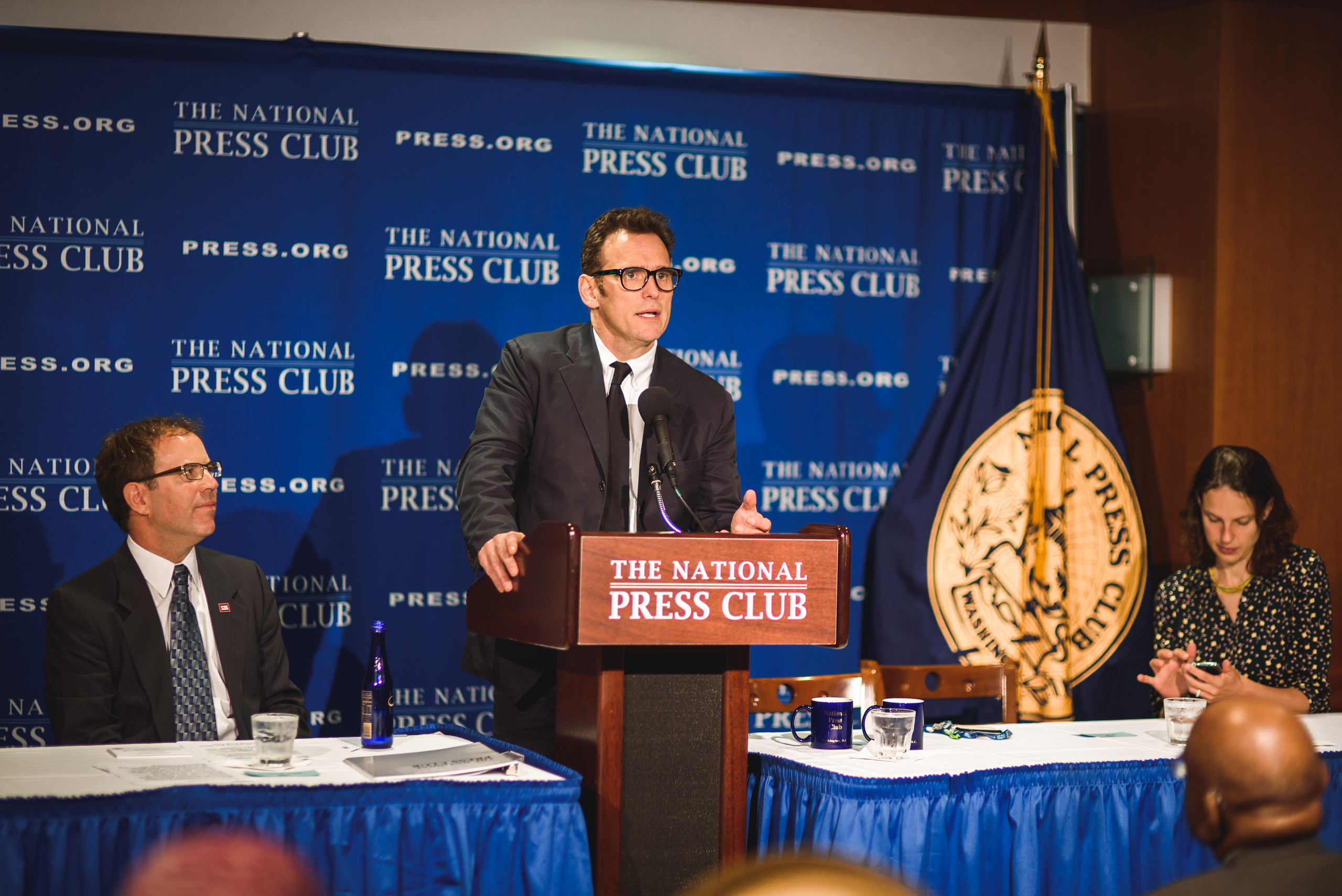 A man in a suit speaks at a podium labeled "The National Press Club" while two other people sit at a table on either side of him. A blue press club banner is in the background.