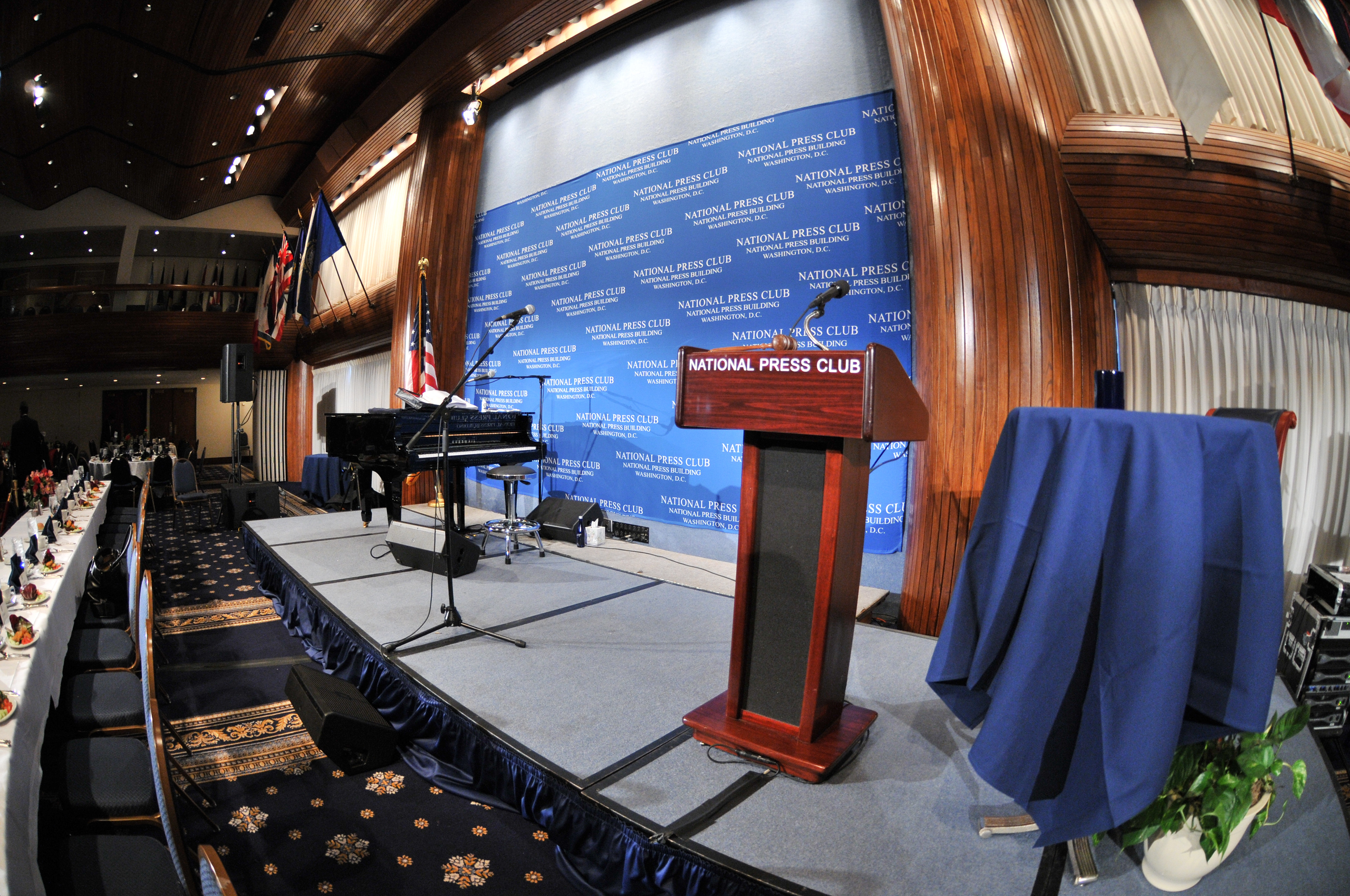 A fisheye view of a stage at the National Press Club with a podium, piano, microphone, and tables set for an event.