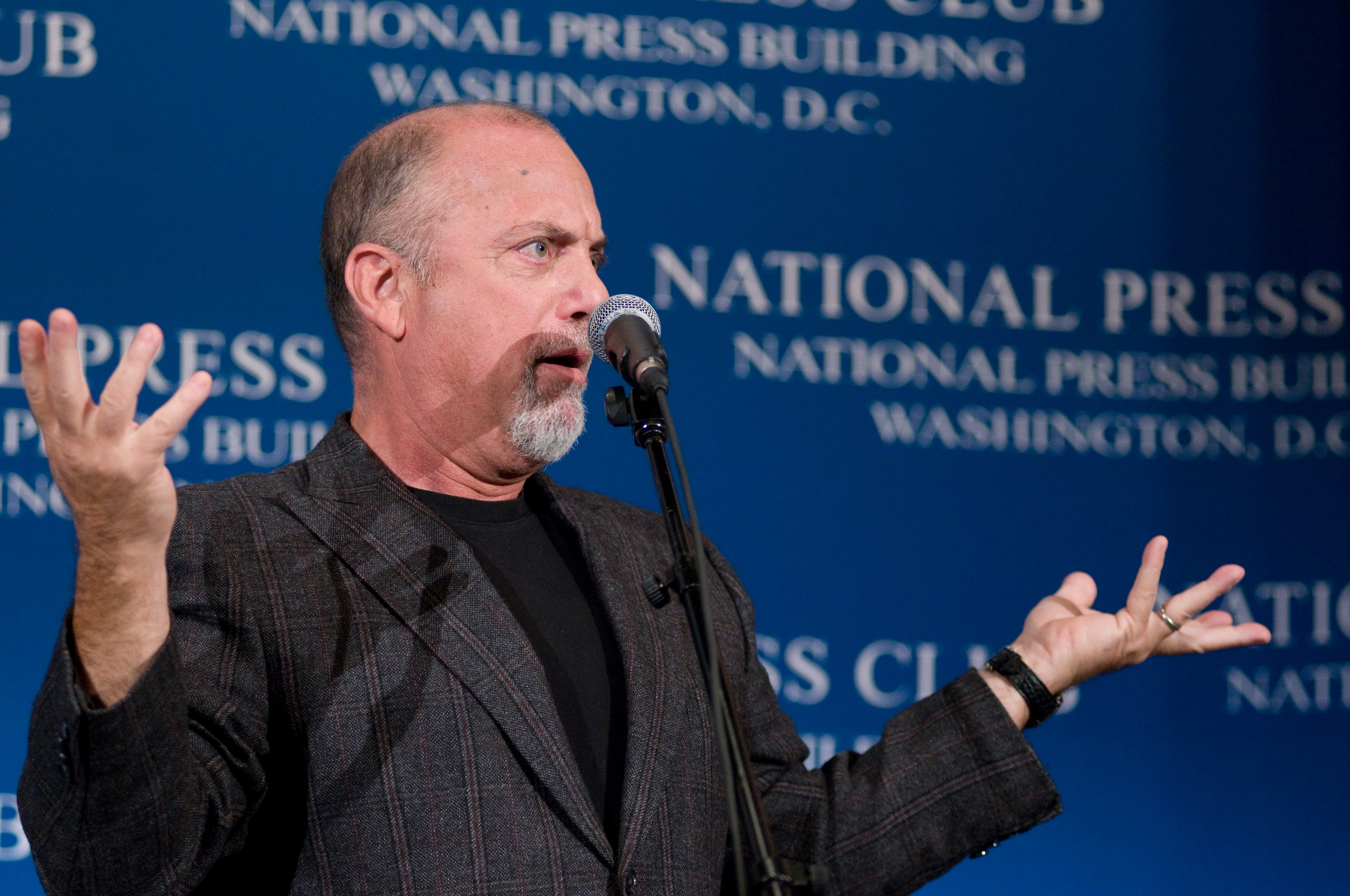 A man speaking into a microphone gestures with both hands, standing in front of a blue backdrop that reads "National Press Club, Washington, D.C.