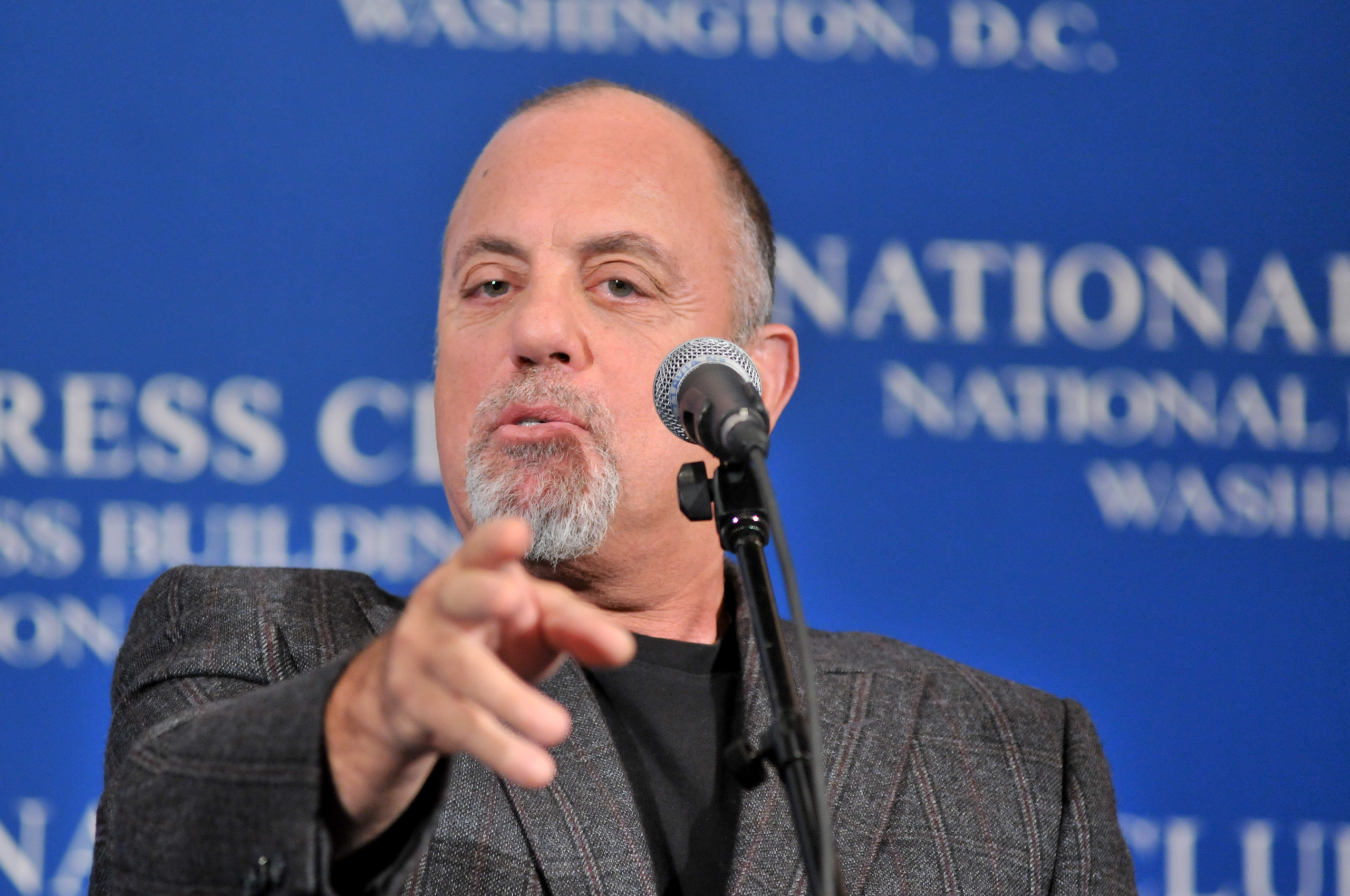 A man with a goatee speaks into a microphone and gestures forward, standing in front of a blue backdrop with white text.