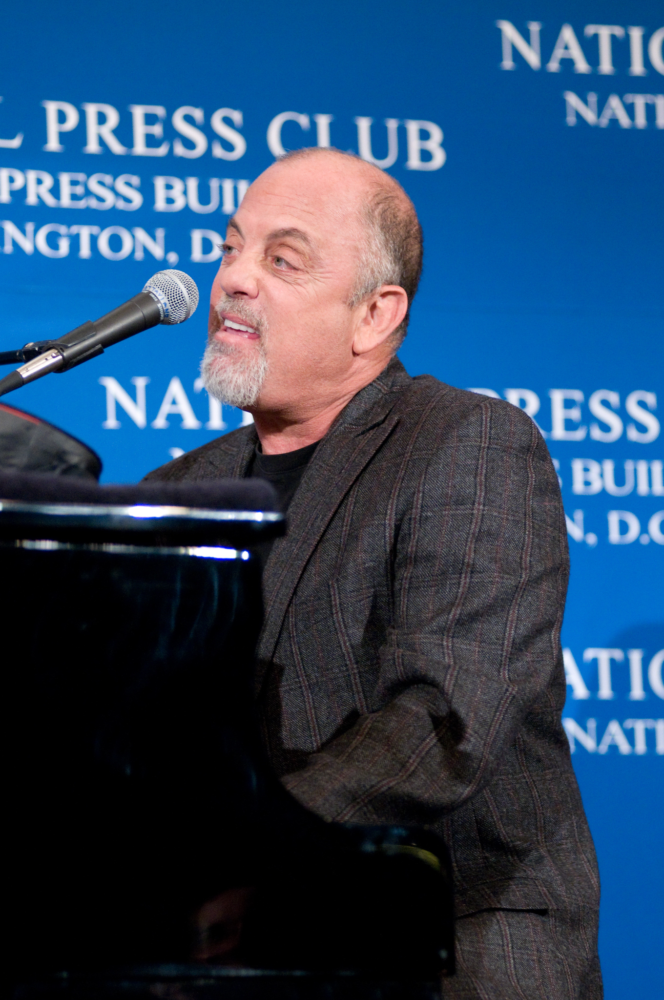 A man with a beard and mustache plays piano and sings into a microphone at a National Press Club event.