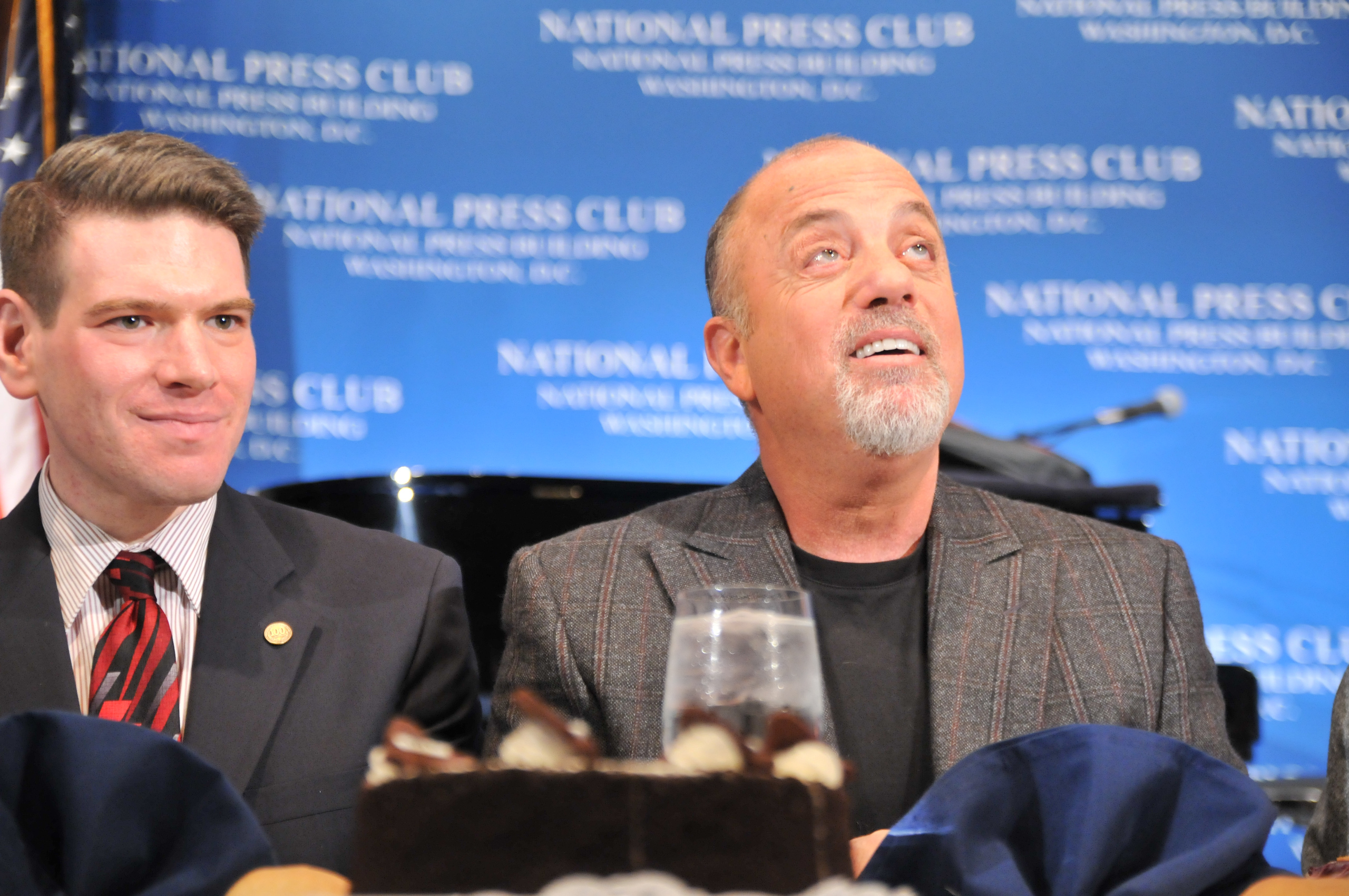 Two men sit at a table with a dessert in front of them, against a blue National Press Club backdrop. One man is wearing a suit and tie, the other a plaid blazer and black shirt.