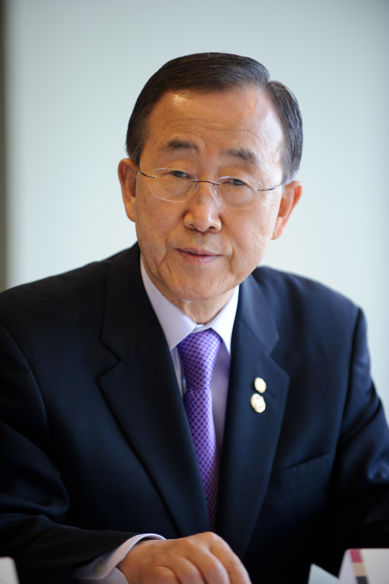 A man in a dark suit and purple tie sits at a table, looking forward. He is wearing glasses and has two pins on his lapel.