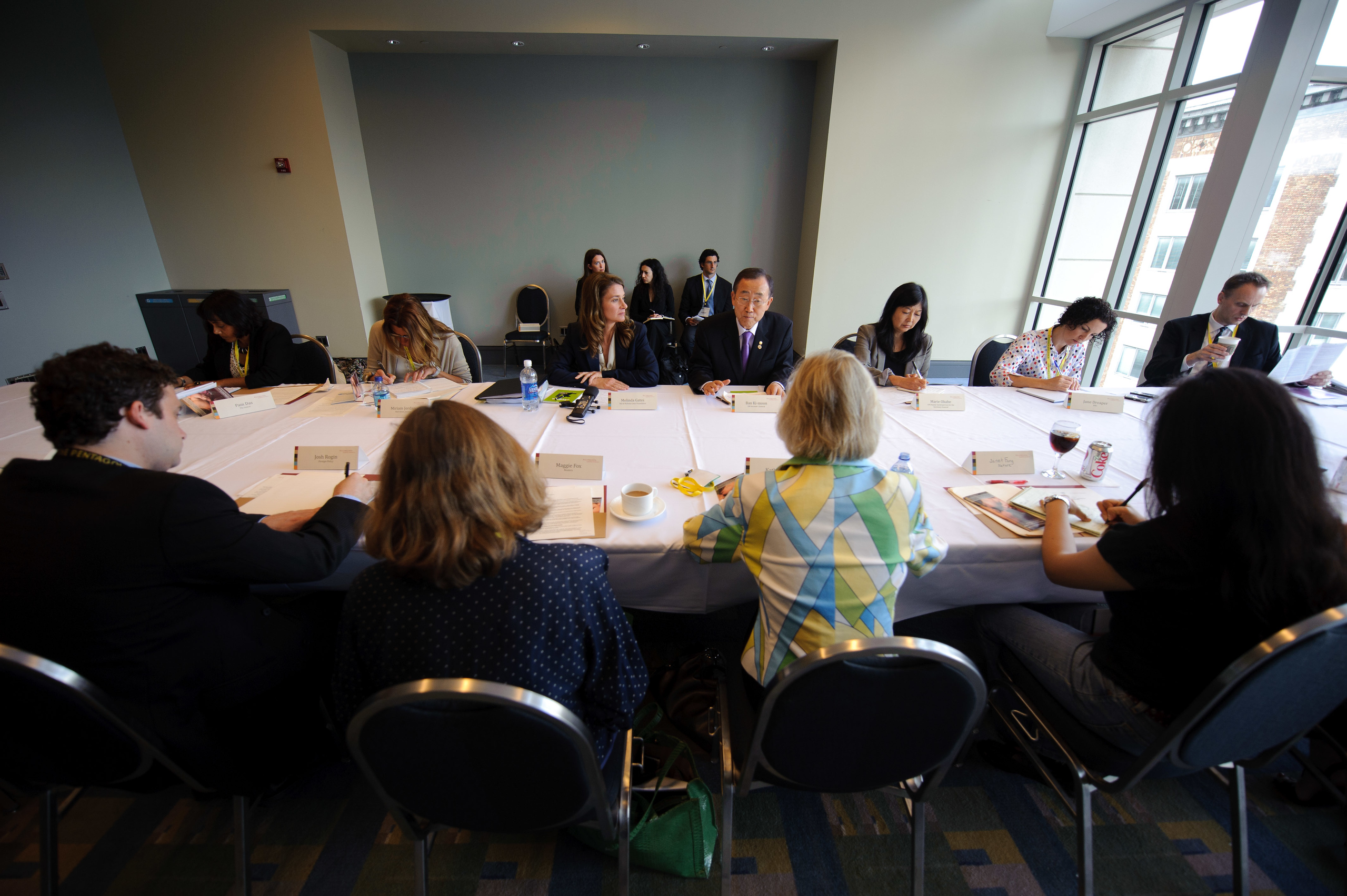 A group of people sit around a large conference table in a meeting room, with papers, drinks, and nameplates in front of them. Several others sit against the back wall.
