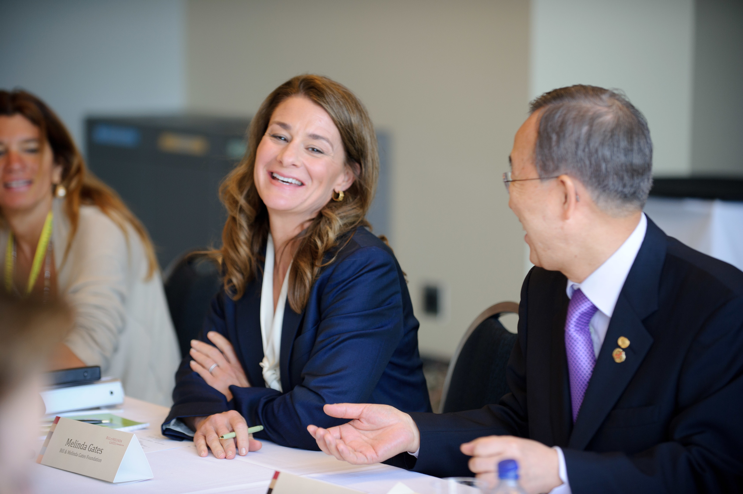 A woman and a man in business attire sit at a table, smiling and conversing during a formal meeting. Name placards and documents are visible on the table.