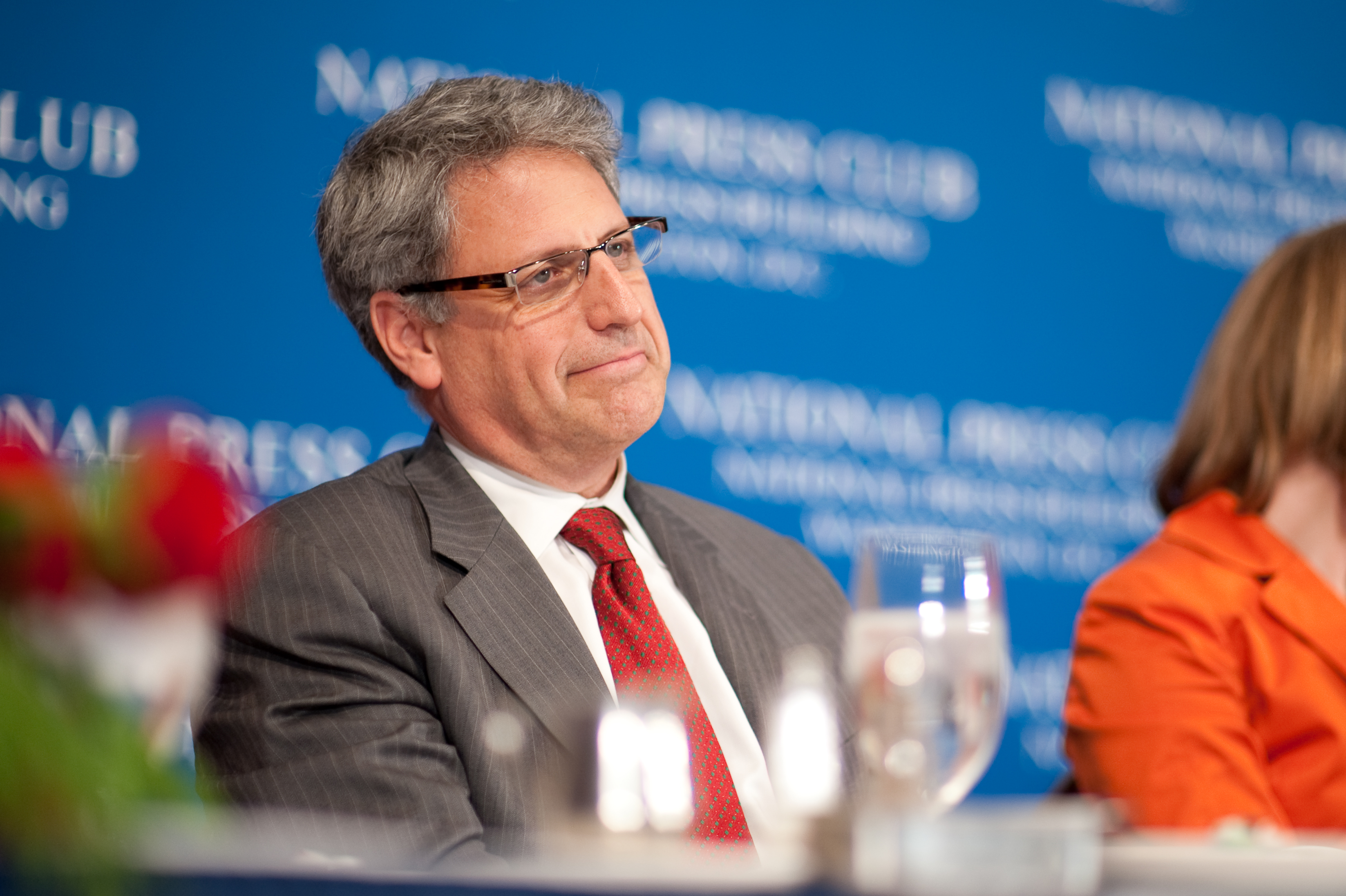 A man in a gray suit and red tie sits at a table with glasses of water in front of him, against a blue National Press Club backdrop.
