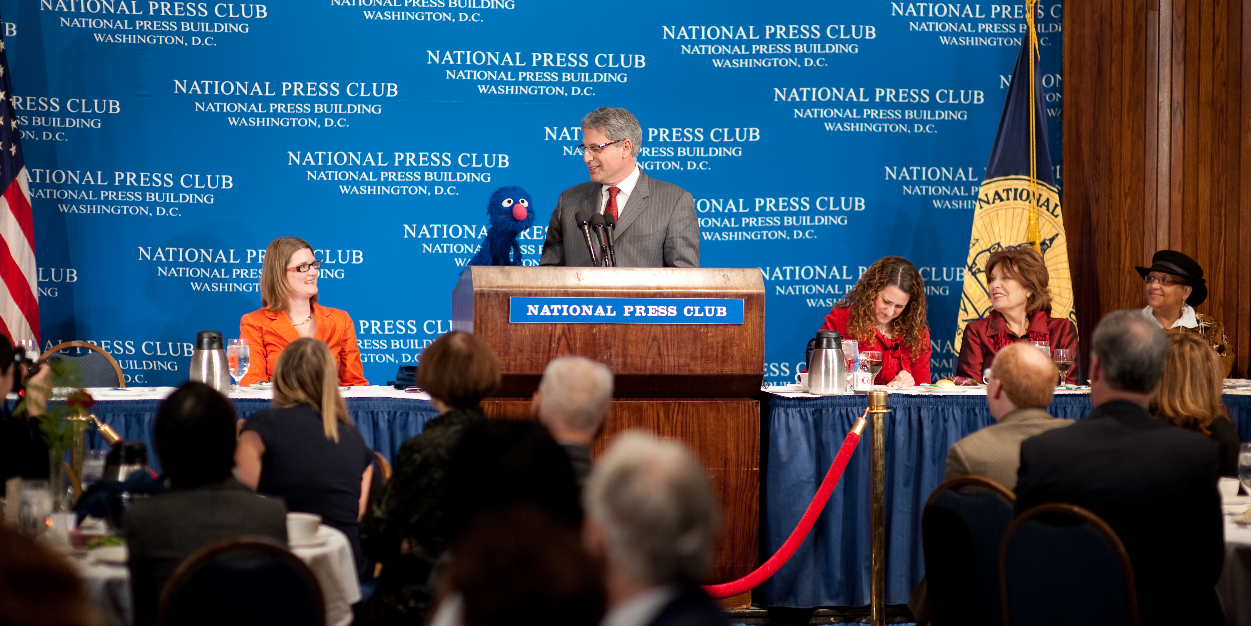 A man speaks at a podium labeled “National Press Club” with a blue background; people are seated at a banquet table on stage and in the audience.