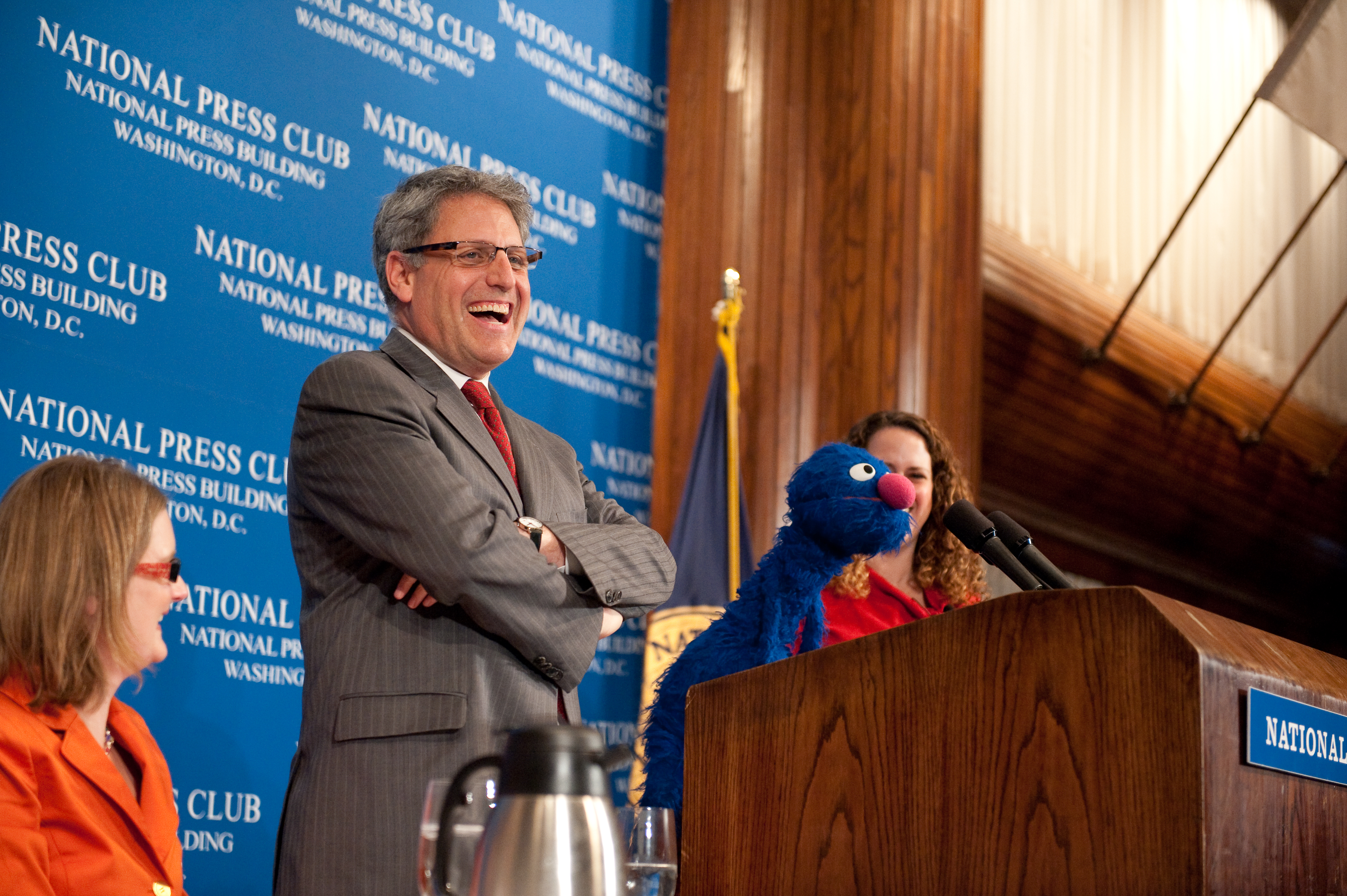 A man stands at a podium smiling with his arms crossed, while a person with a Grover puppet speaks into microphones at a National Press Club event.