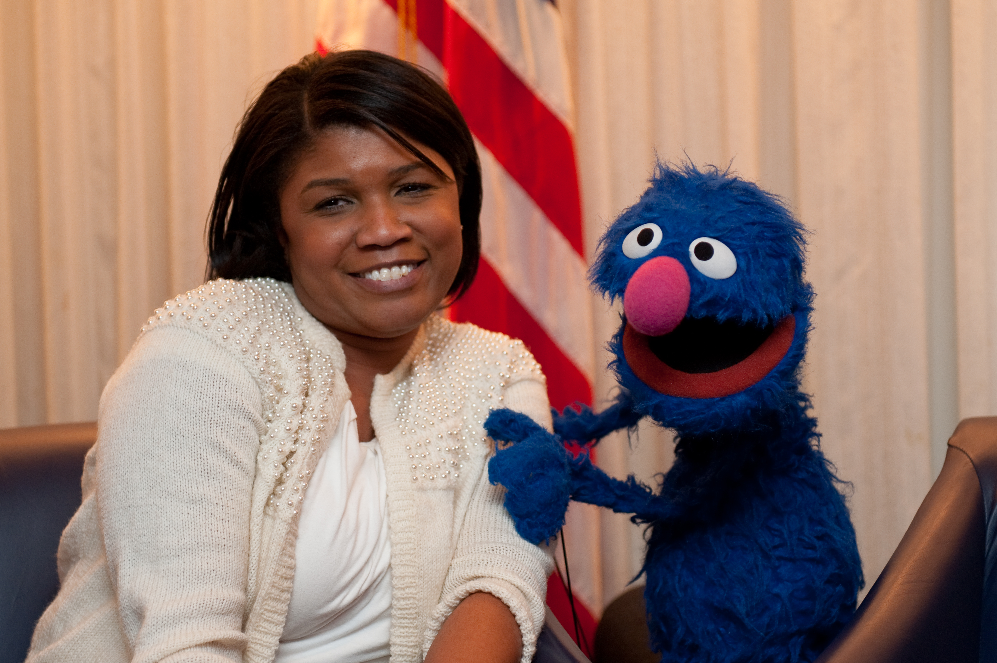 A woman sits and smiles next to the blue puppet character Grover from Sesame Street, with part of an American flag visible in the background.