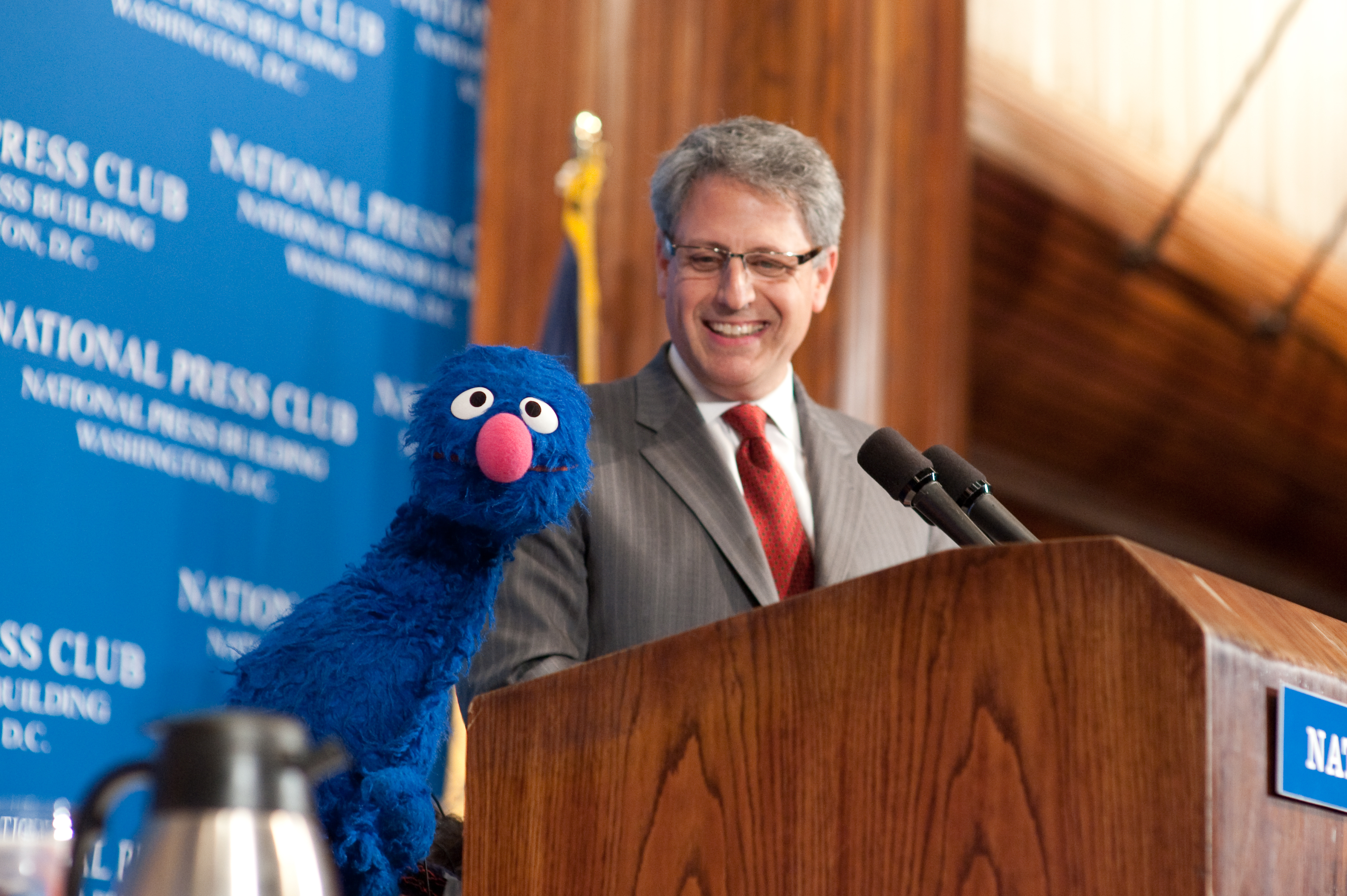 A man in a suit stands at a podium with microphones, smiling next to Grover, a blue Muppet character, at a National Press Club event.
