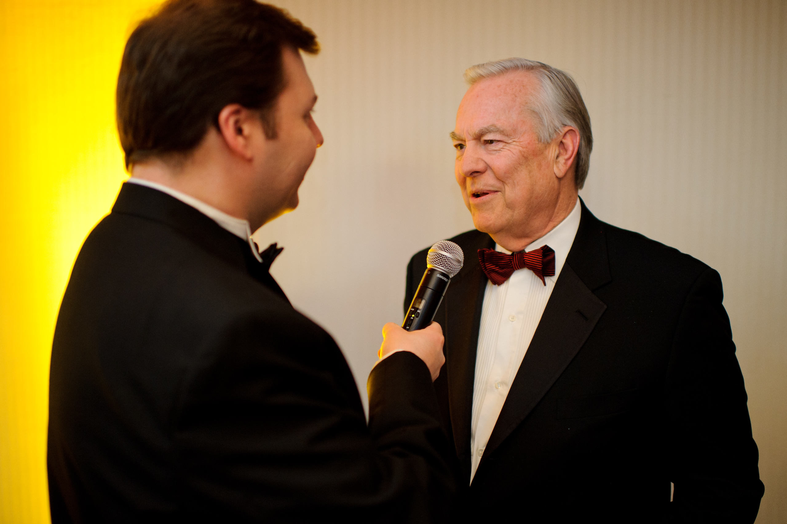 A man in a tuxedo interviews another man in a tuxedo and red bow tie, holding a microphone to him against a plain background.