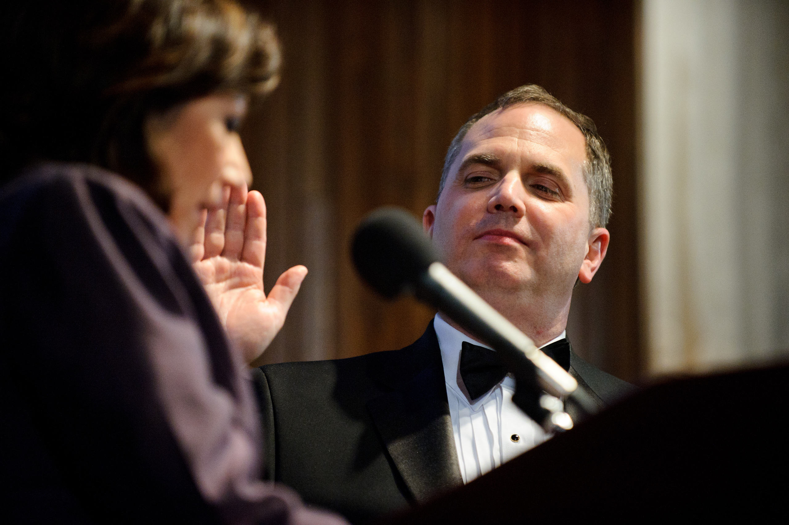 A man in a tuxedo raises his right hand, appearing to take an oath, while a woman in the foreground speaks into a microphone.