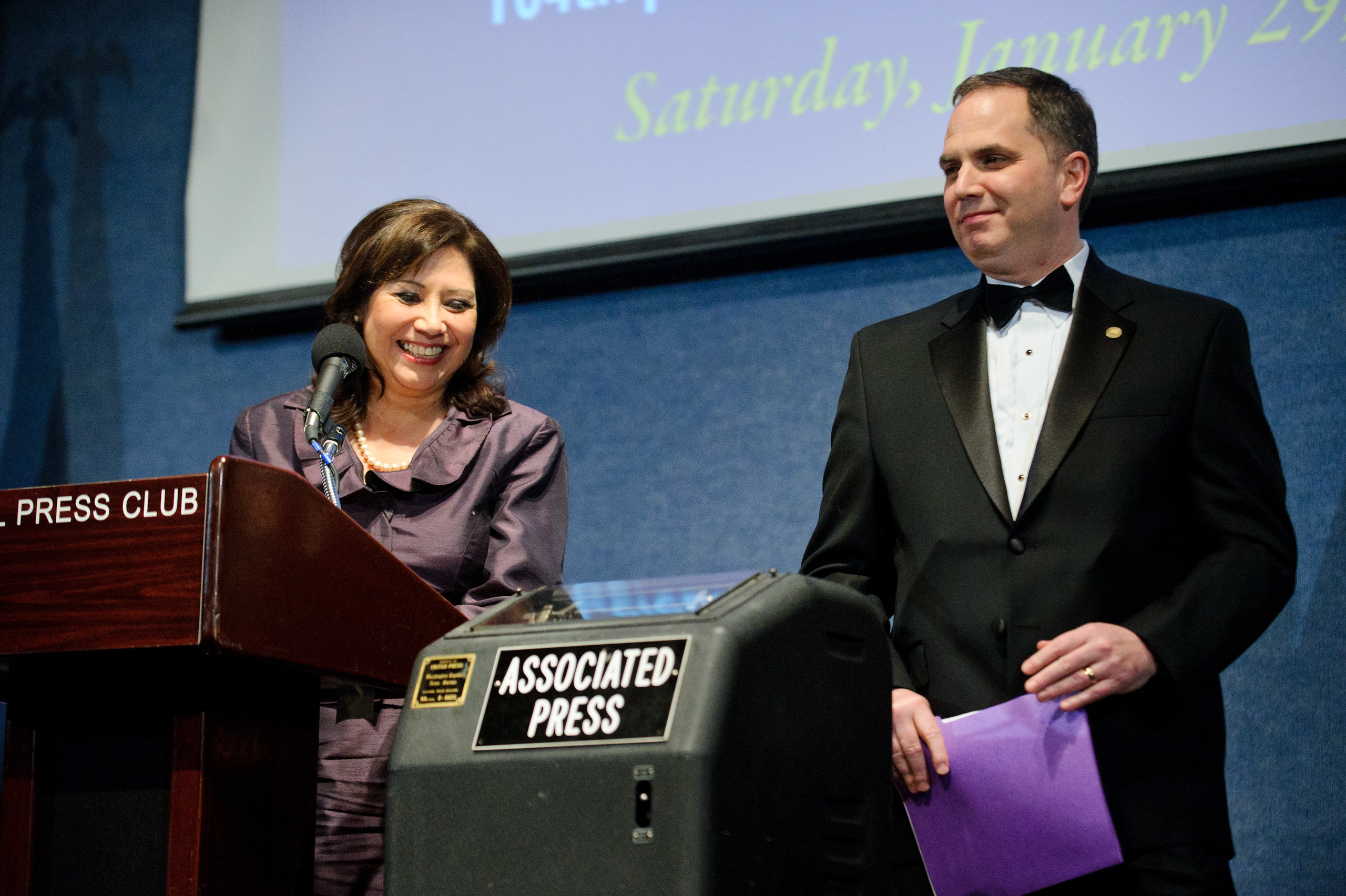 A woman speaks at a podium labeled "PRESS CLUB" while a man in a tuxedo stands beside her, both near an "ASSOCIATED PRESS" sign.