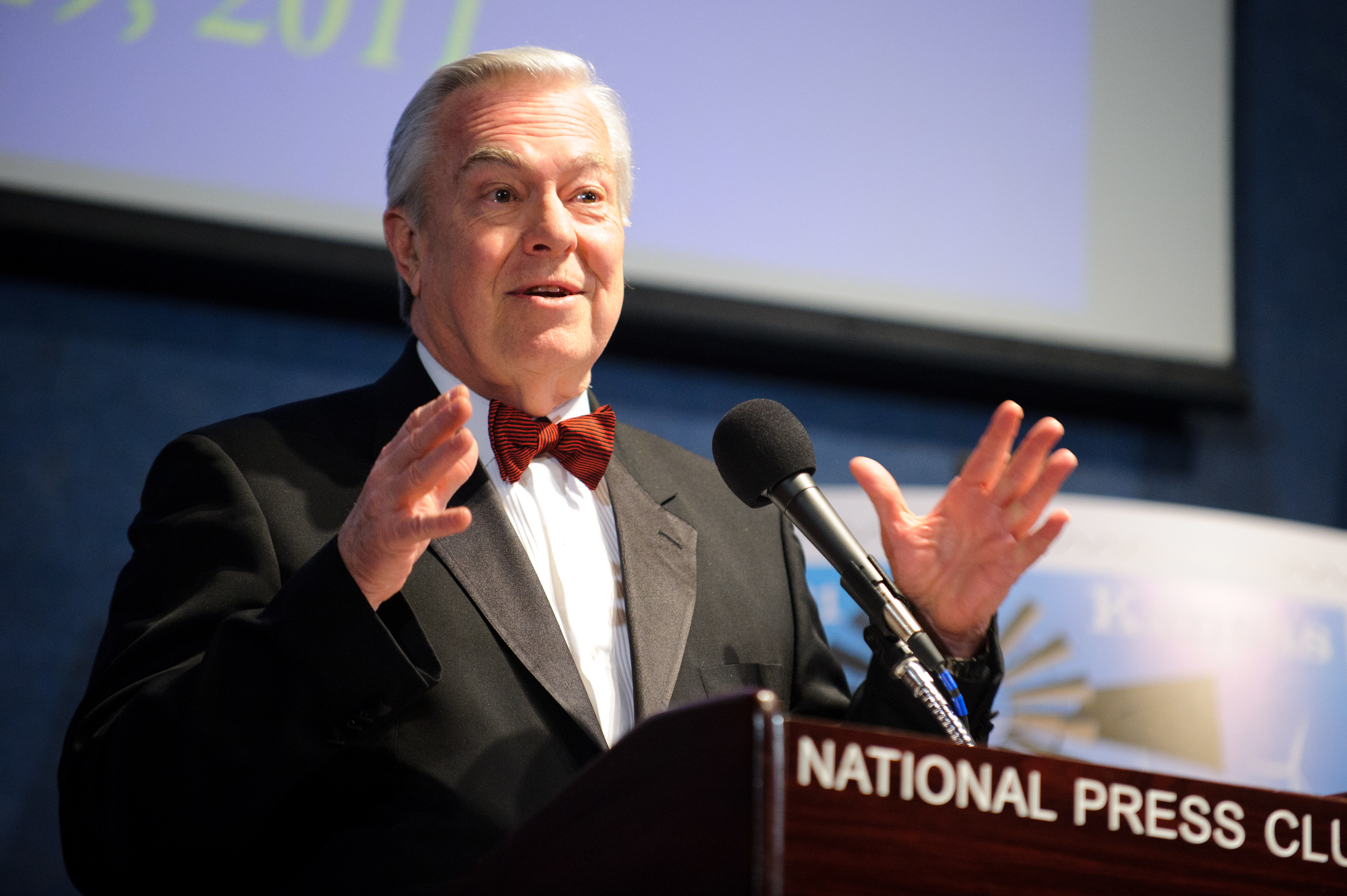An older man in a suit and bow tie speaks at a podium labeled "National Press Club," gesturing with his hands, with a microphone in front of him.