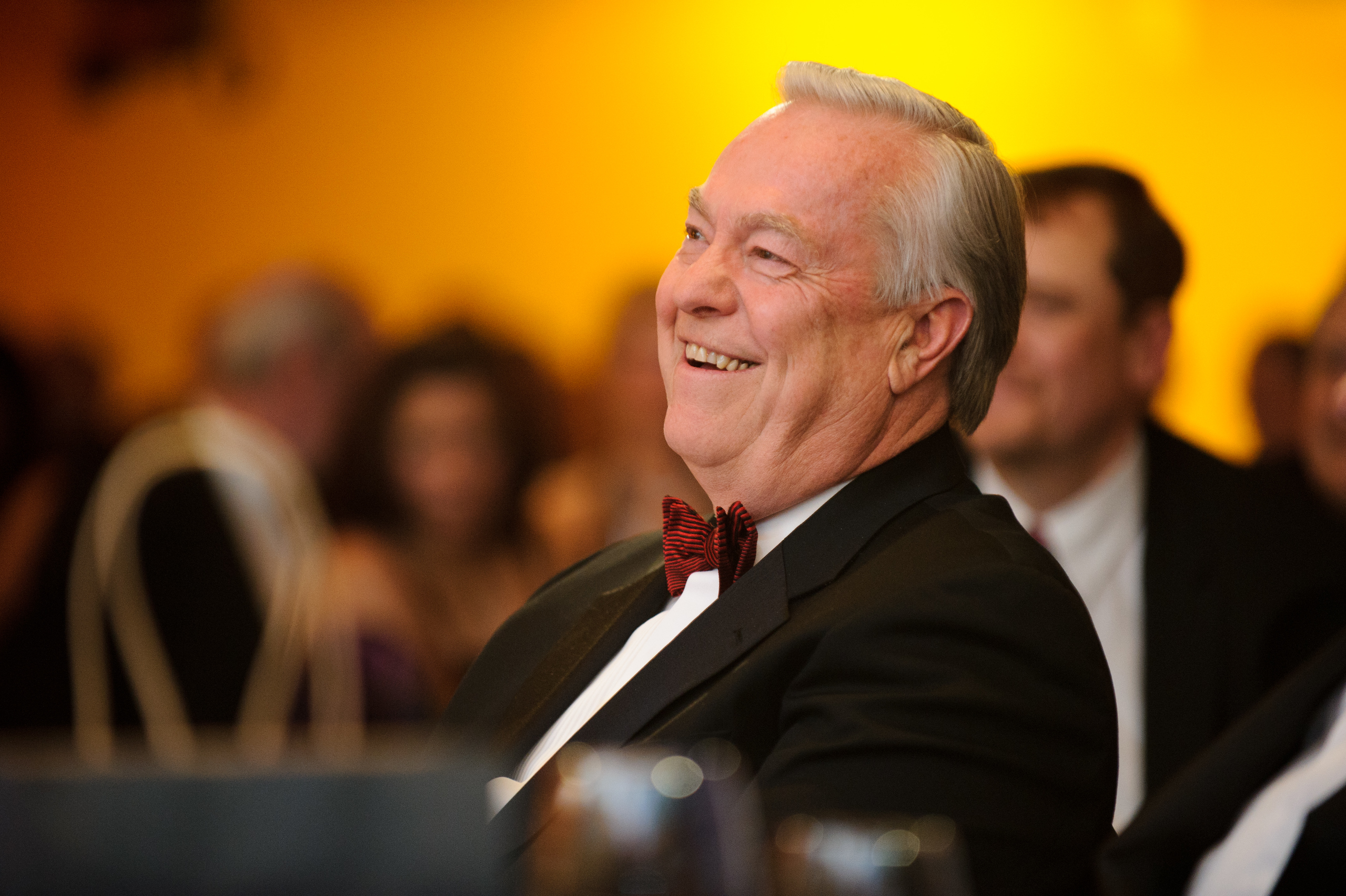 An older man in a tuxedo and red bow tie smiles while seated at a formal event with a blurred crowd in the background.