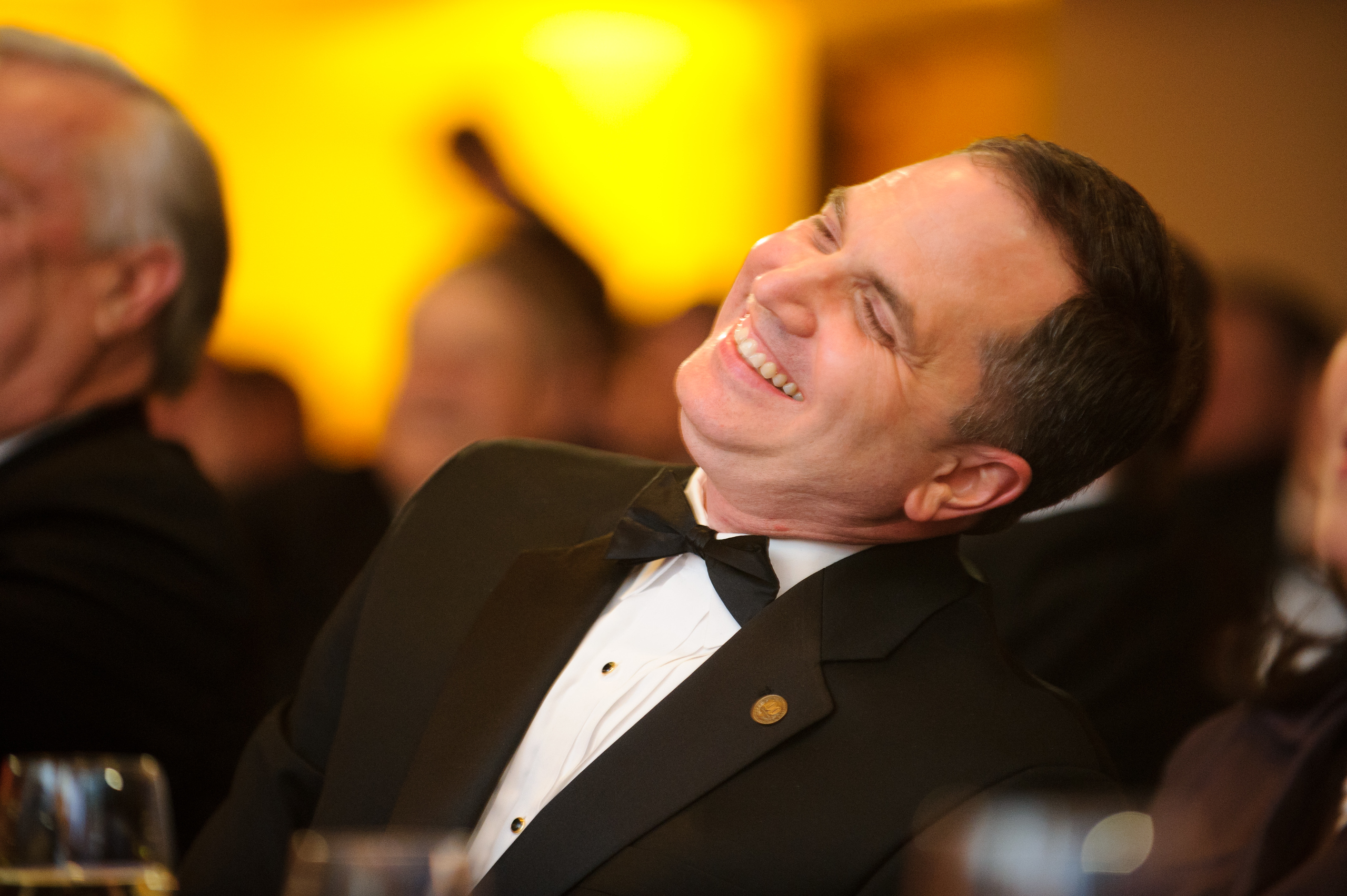 A man in a tuxedo leans back and laughs at a formal event, with blurred attendees and warm lighting in the background.