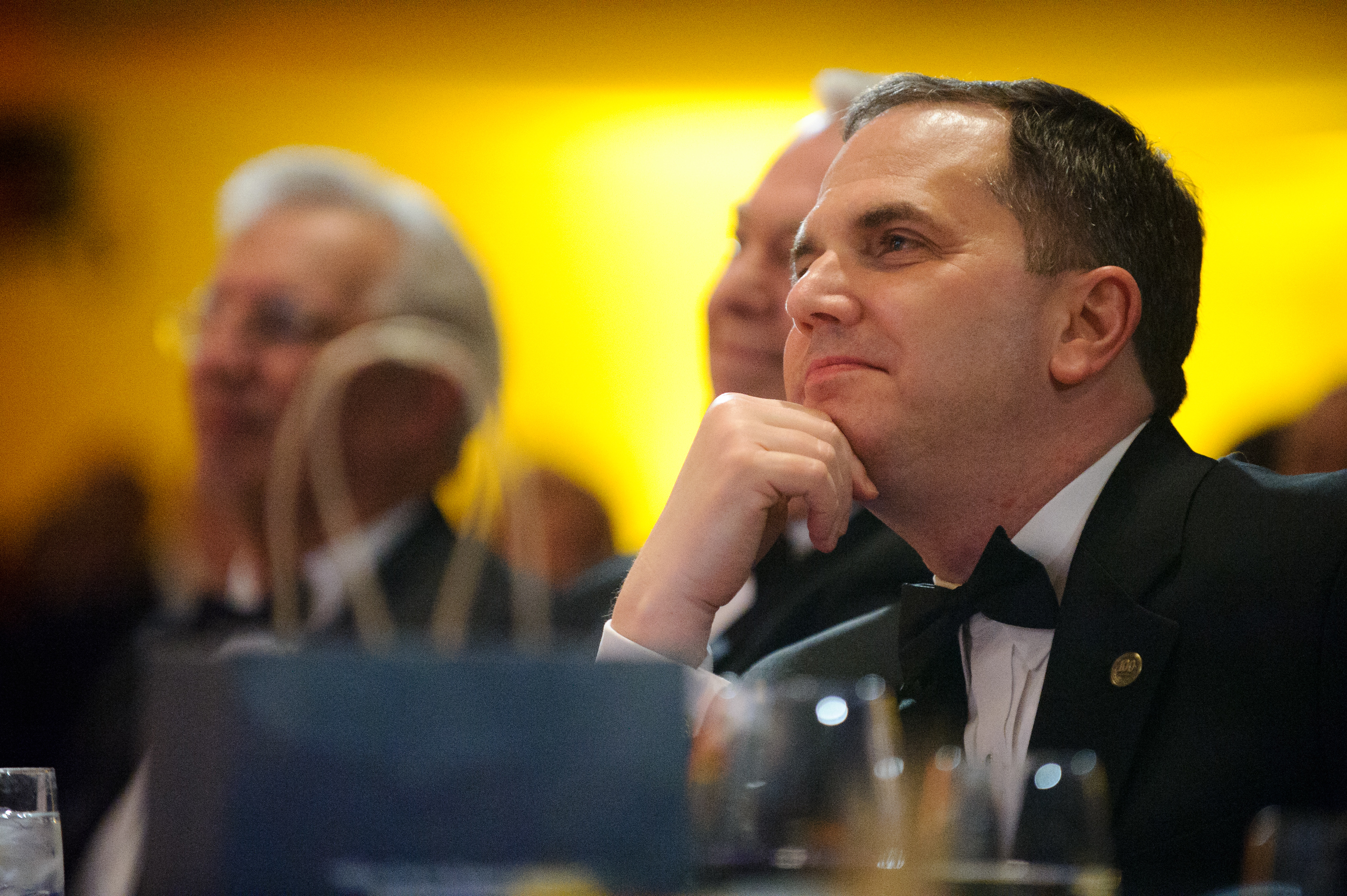 A man in a black tuxedo sits at a table, resting his chin on his hand and looking ahead, with other people seated in the blurred background.