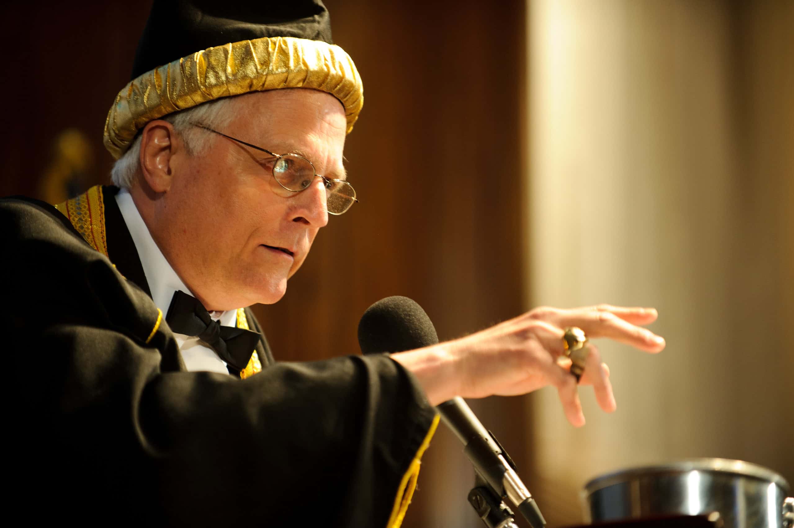 An older man in ceremonial robes and hat gestures while speaking into a microphone at a podium.