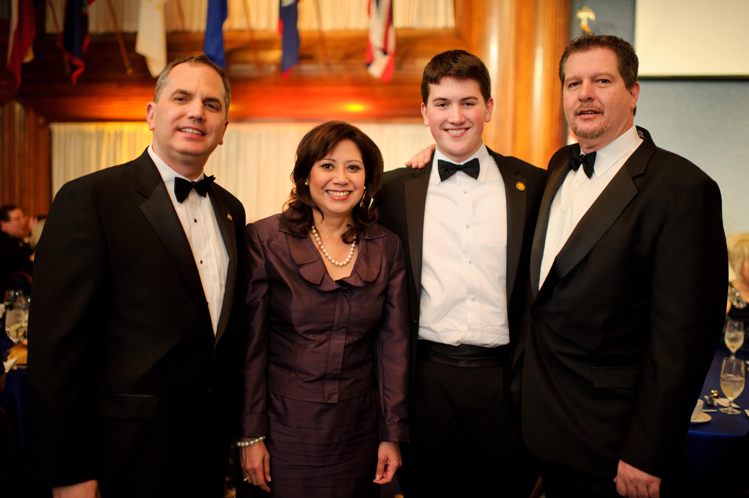 Four people dressed formally stand together at an indoor event; three men in black tuxedos and one woman in a purple dress.