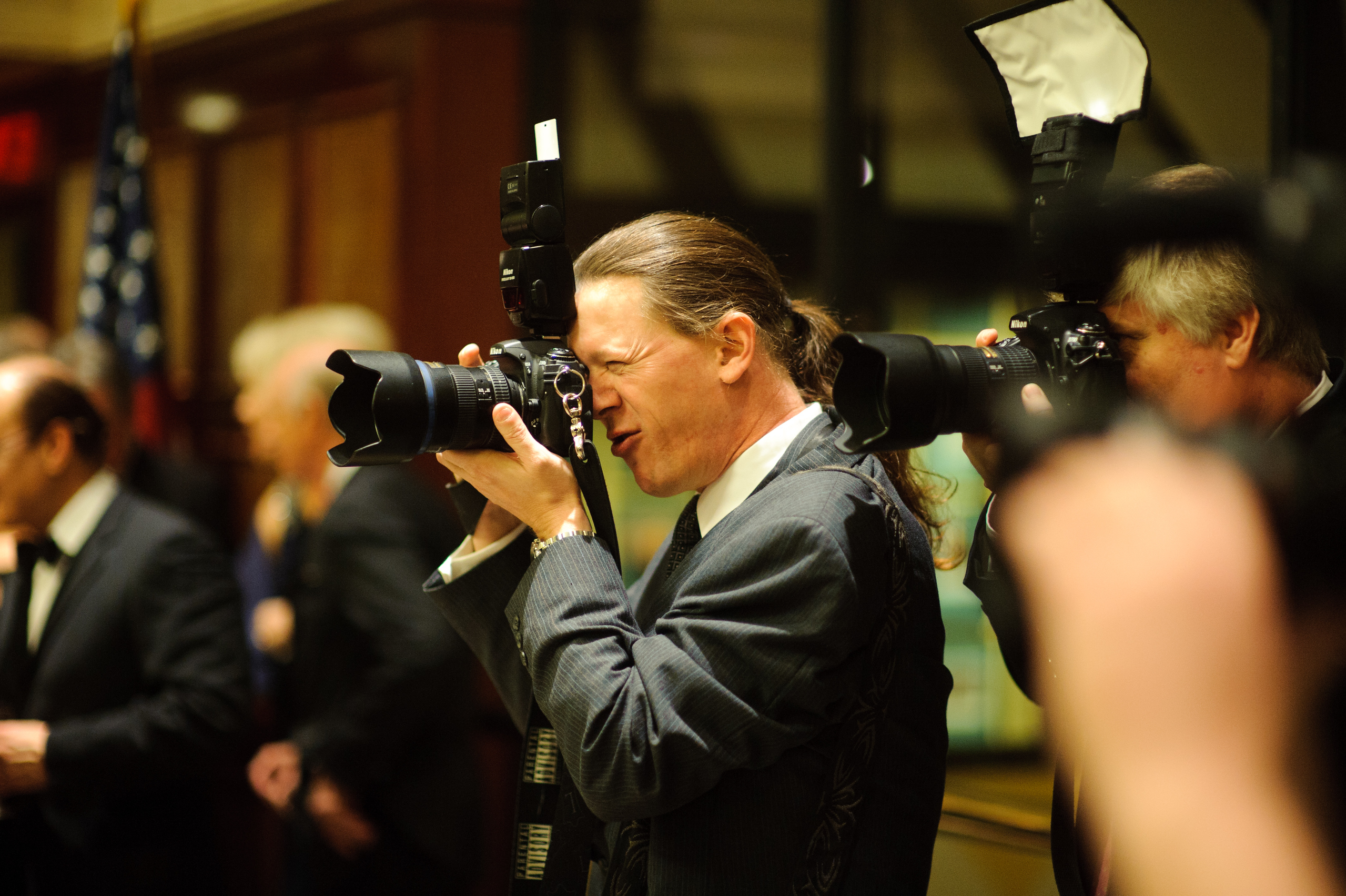 A man in a suit with long hair tied back is taking a photo with a professional camera at an indoor event. Other people and photographers are visible in the background.
