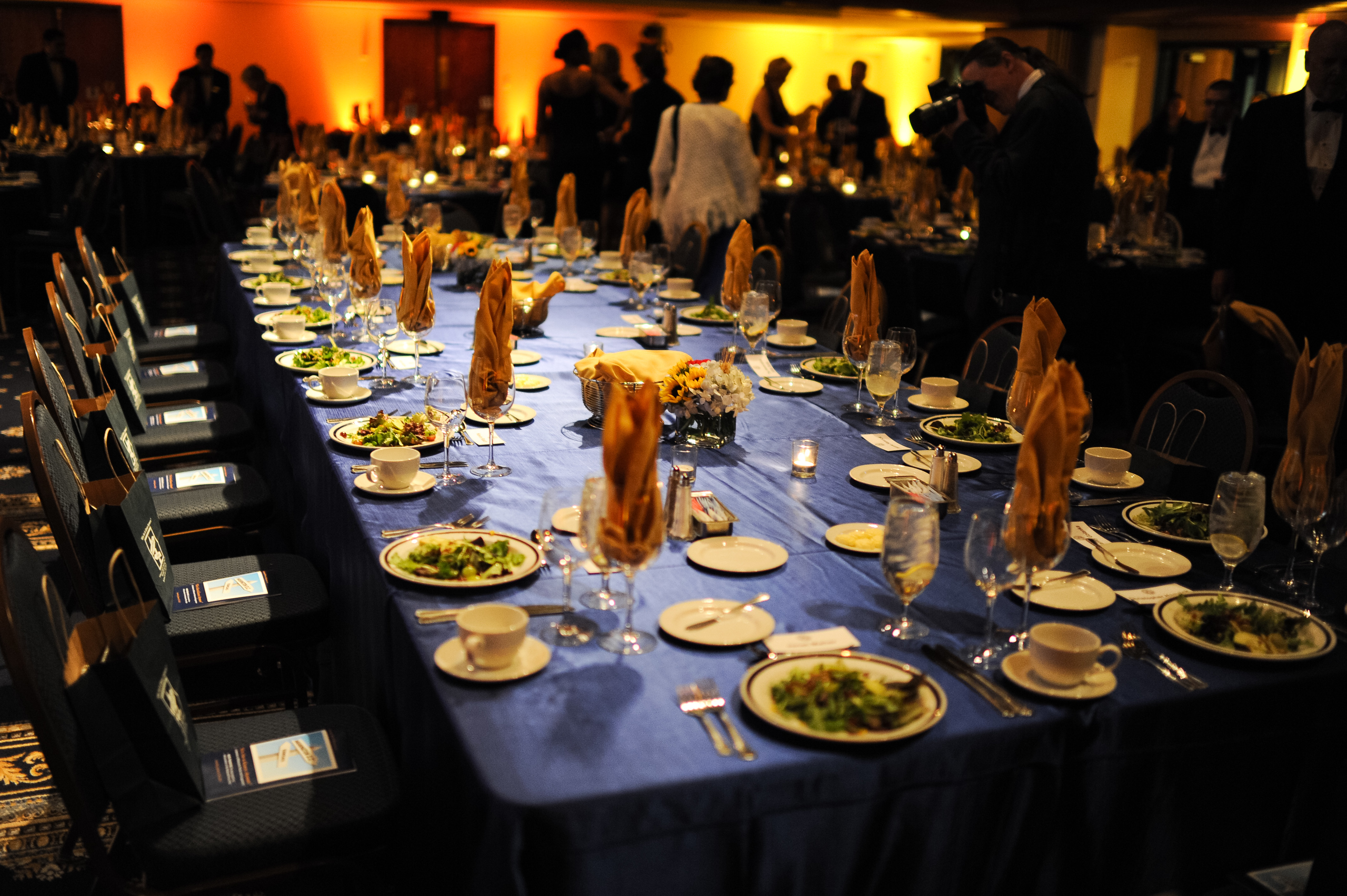 A banquet table covered with a blue tablecloth is set with salads, napkins, cups, and glasses at a formal event, with people standing and mingling in the background.