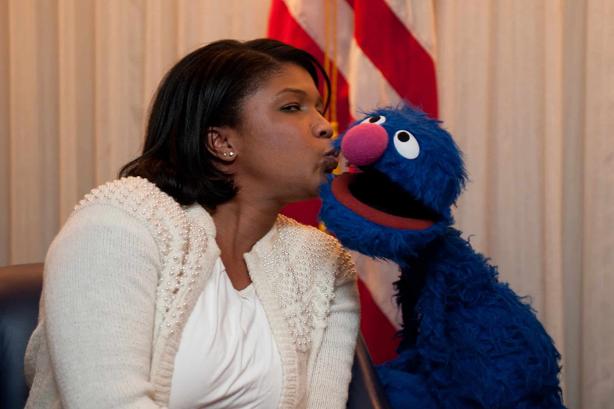 A woman sits beside Grover Monster the Muppet, who is leaning in close to her face in this epic portrait. An American flag is visible in the background.