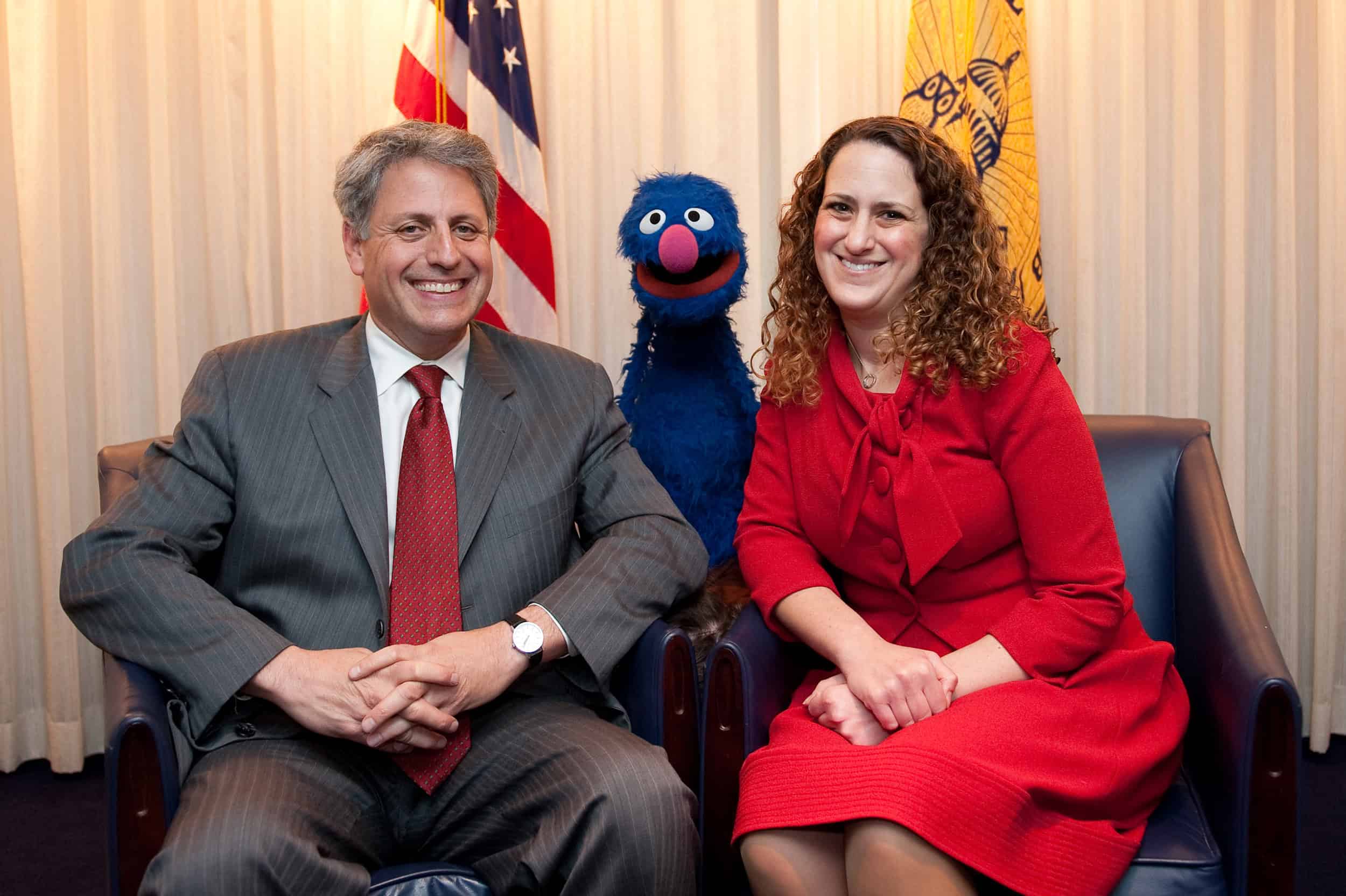 A man in a gray suit, a woman in a red dress, and Grover Monster sit together on chairs in front of U.S. flags, creating an epic portrait of camaraderie and color.