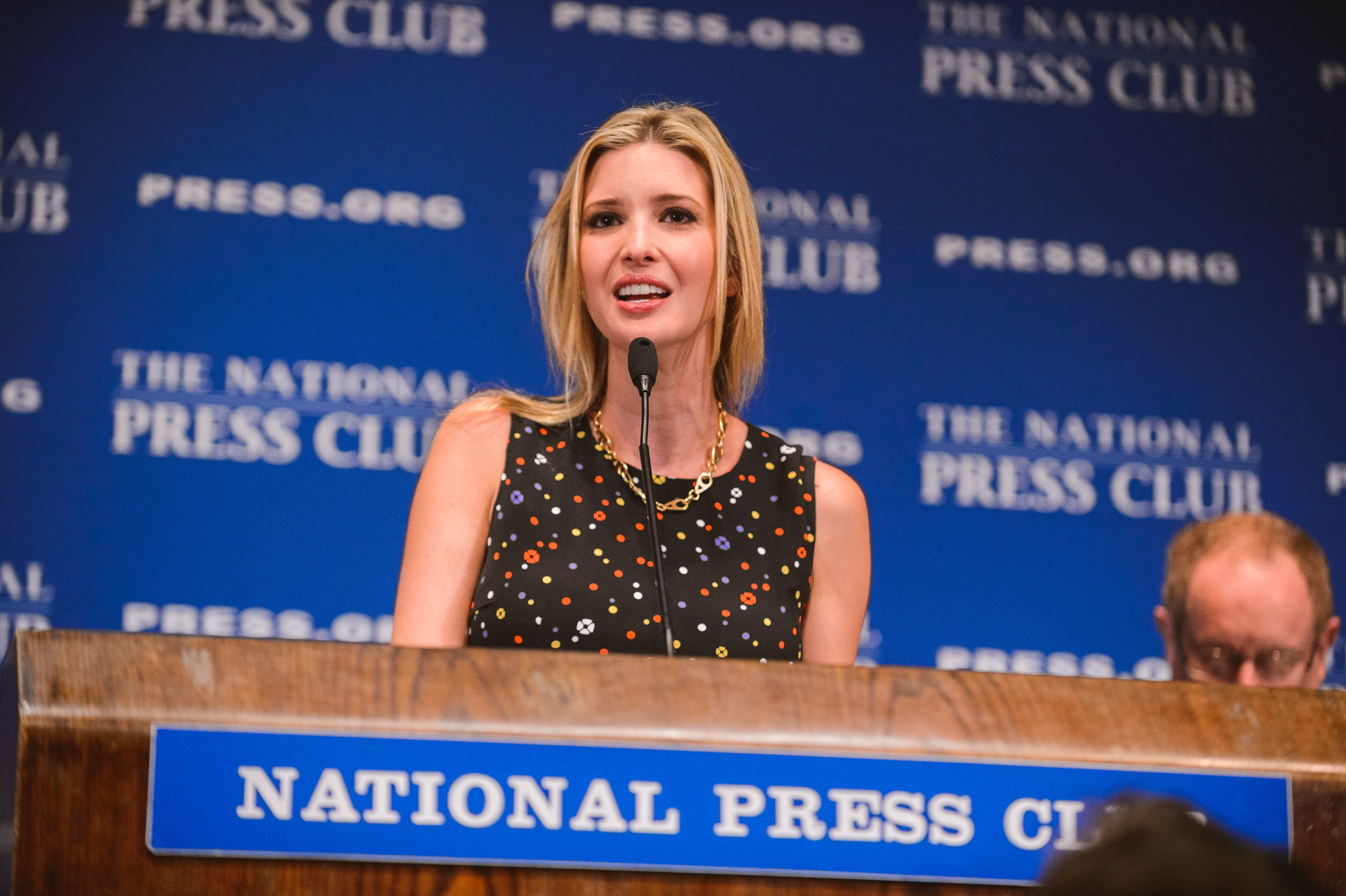 A woman speaks at a podium labeled "National Press Club" with a blue backdrop displaying "The National Press Club" and "Press.org" logos.