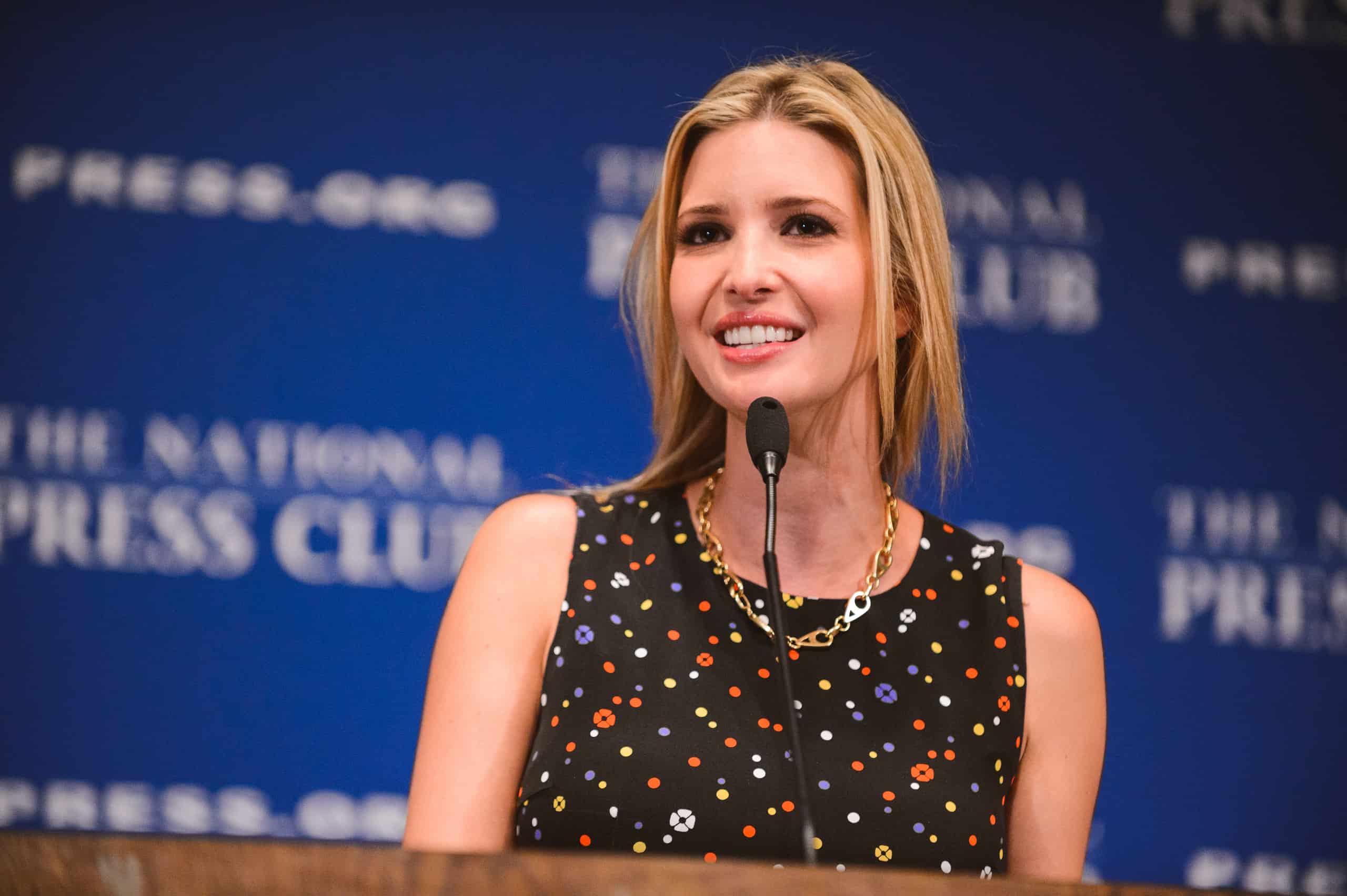 A woman in a black sleeveless dress speaks at a podium with a microphone in front of a blue background that reads "The National Press Club.
