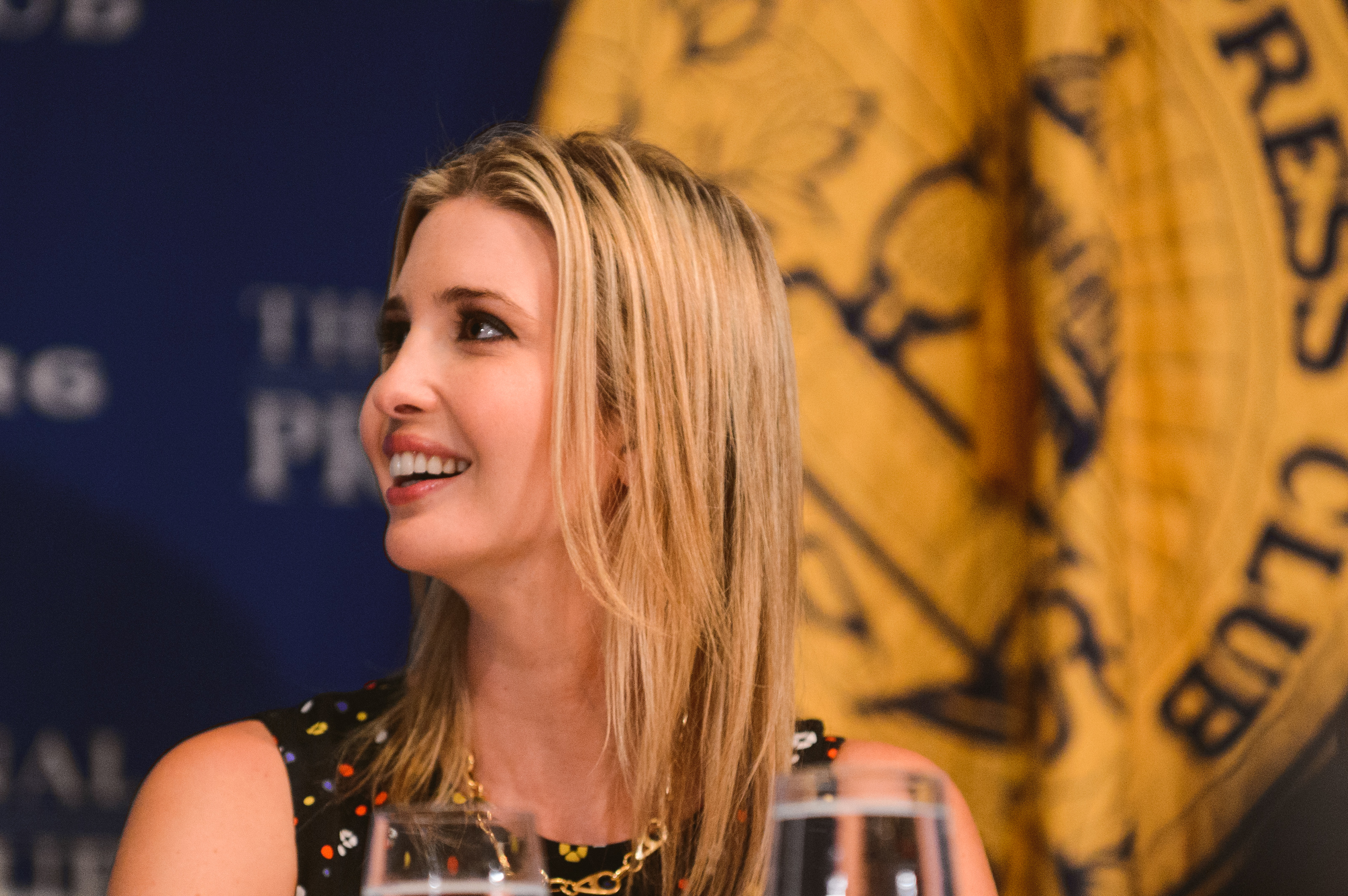A woman with long blonde hair and a black dress sits at a table, smiling and looking to the side, with a large gold emblem in the background.
