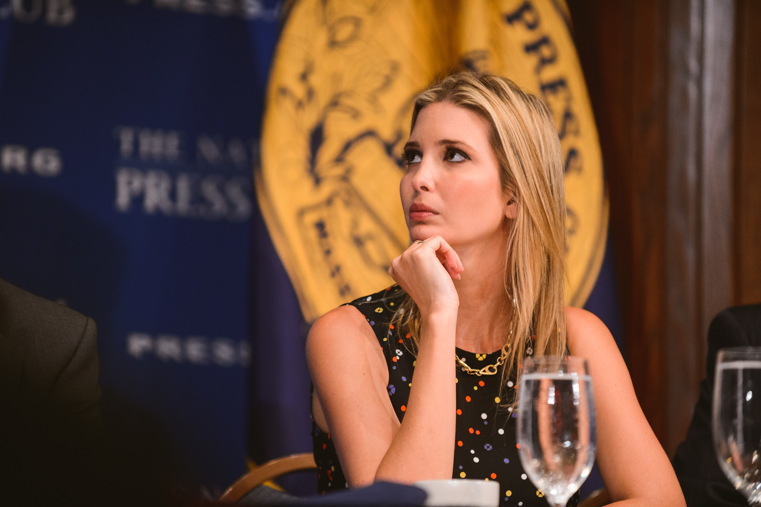 A woman with blonde hair sits at a table with a glass of water, resting her chin on her hand, in front of blue and yellow press club banners.