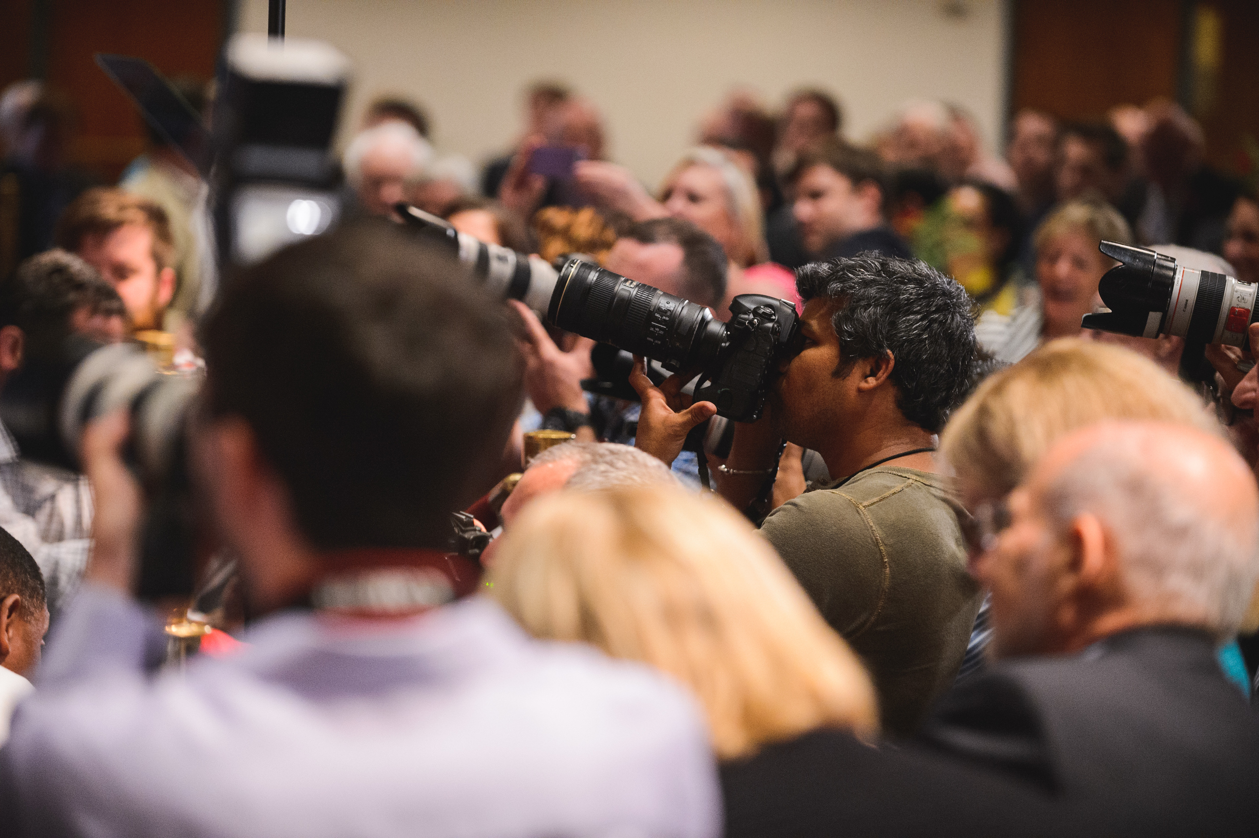 A group of photographers with cameras and long lenses take photos in a crowded indoor event with many people in the background.
