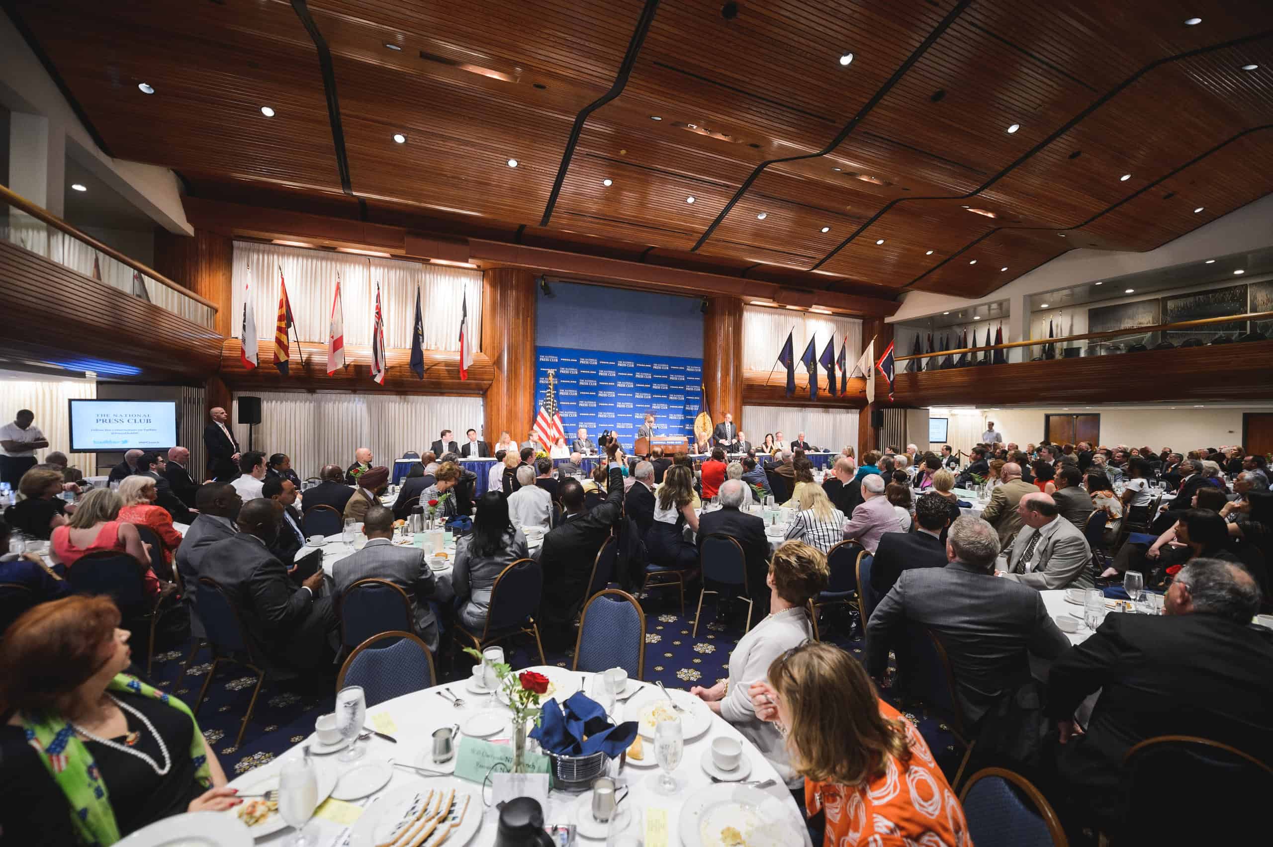 Large conference room filled with people seated at round tables, facing a stage with speakers, flags, and a blue backdrop.
