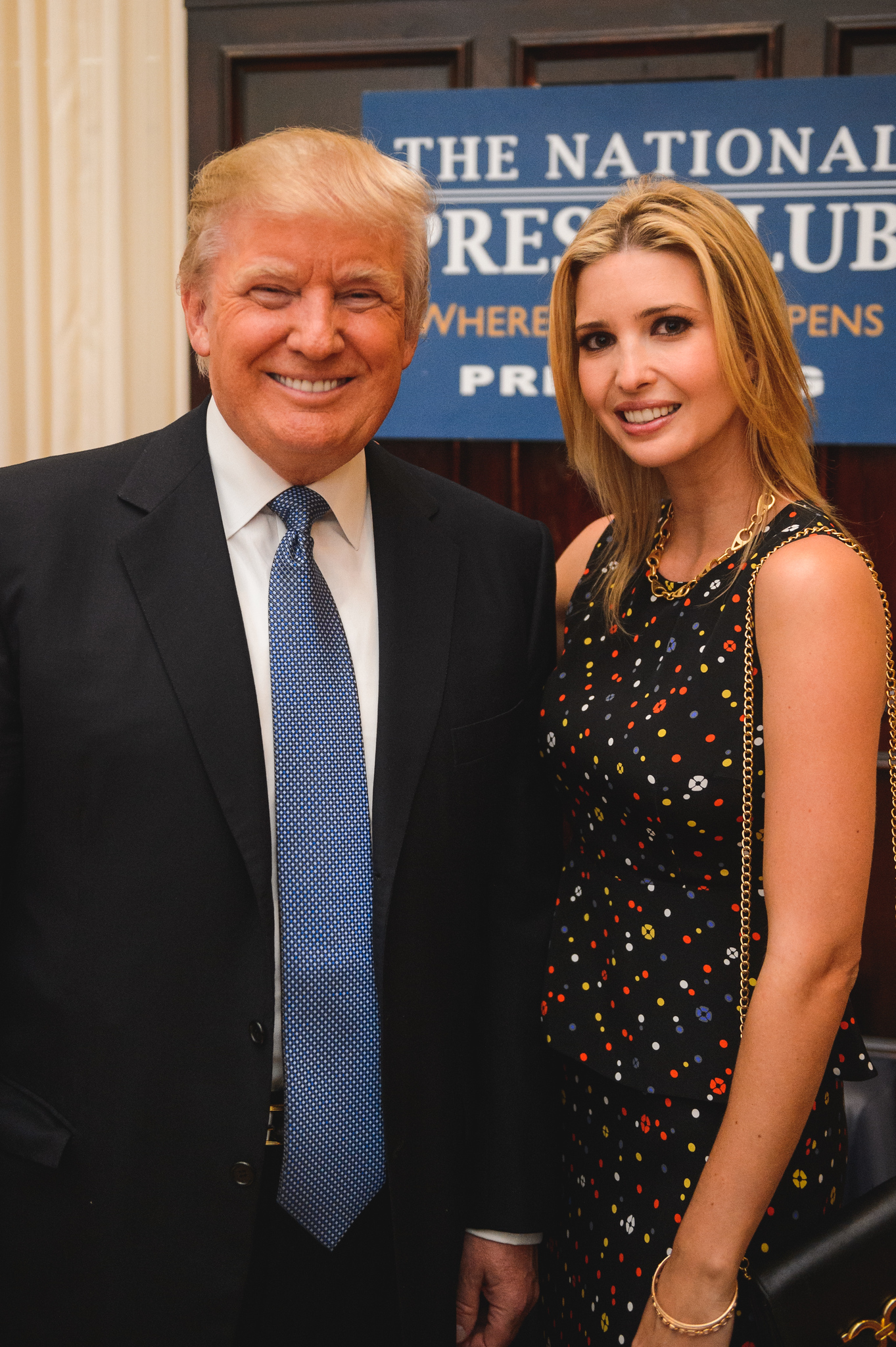 Two people in formal attire stand and smile in front of a sign that reads "The National Press Club.