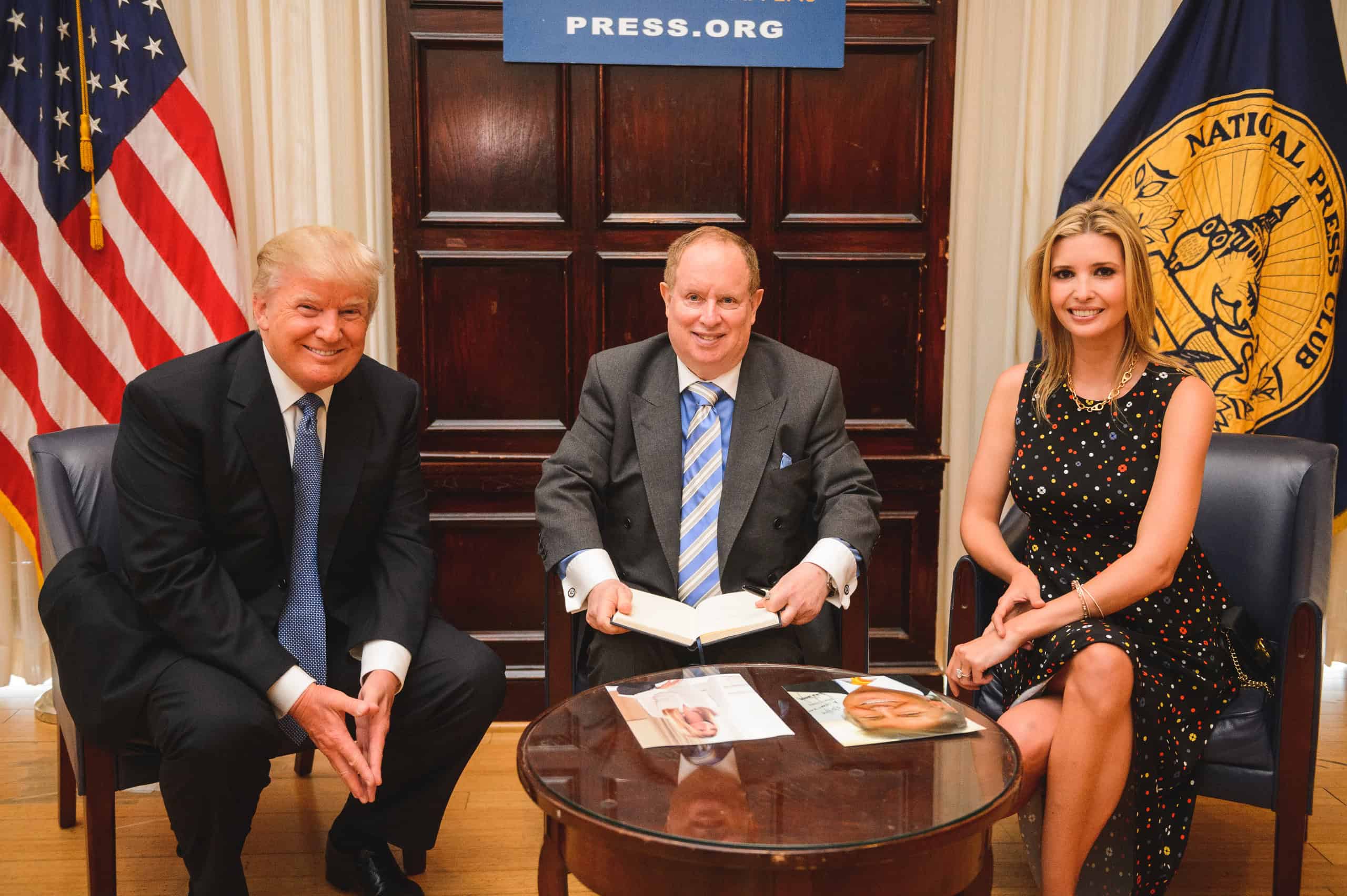 Three people pose for a photo in an office setting; two sit on either side of a man holding an open book, with magazines on the table in front of them and flags in the background.