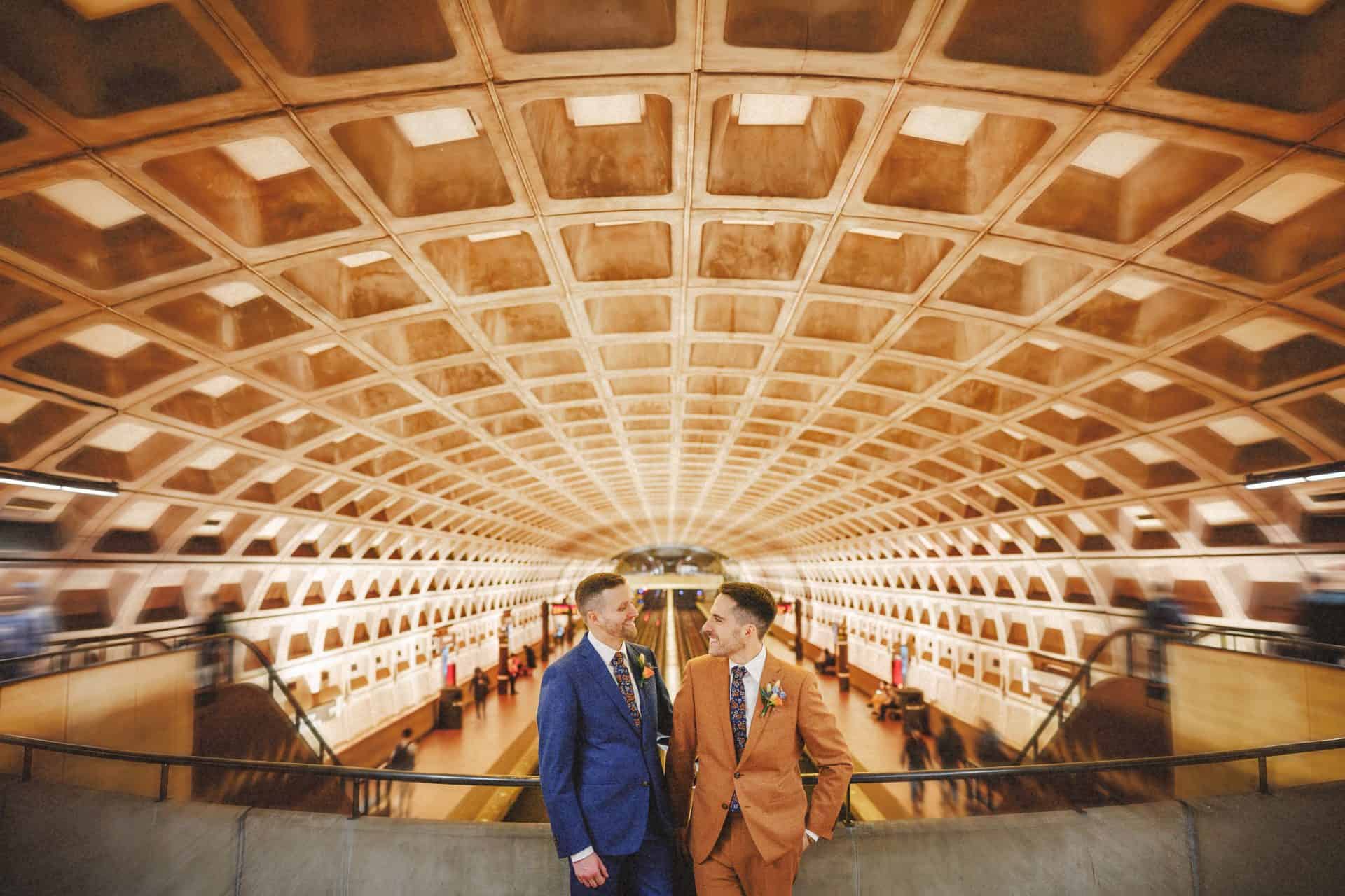 Two grooms in blue and rust suits posing under the coffered ceiling of a Washington DC Metro station