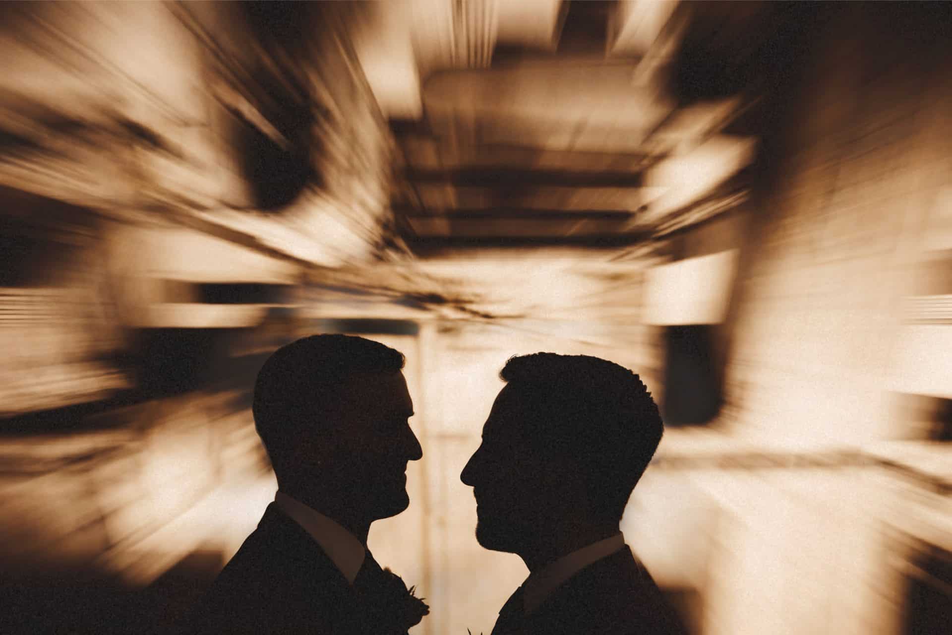 Two grooms in rust and blue suits holding hands on a bench in front of the book walls at Planet Word in DC