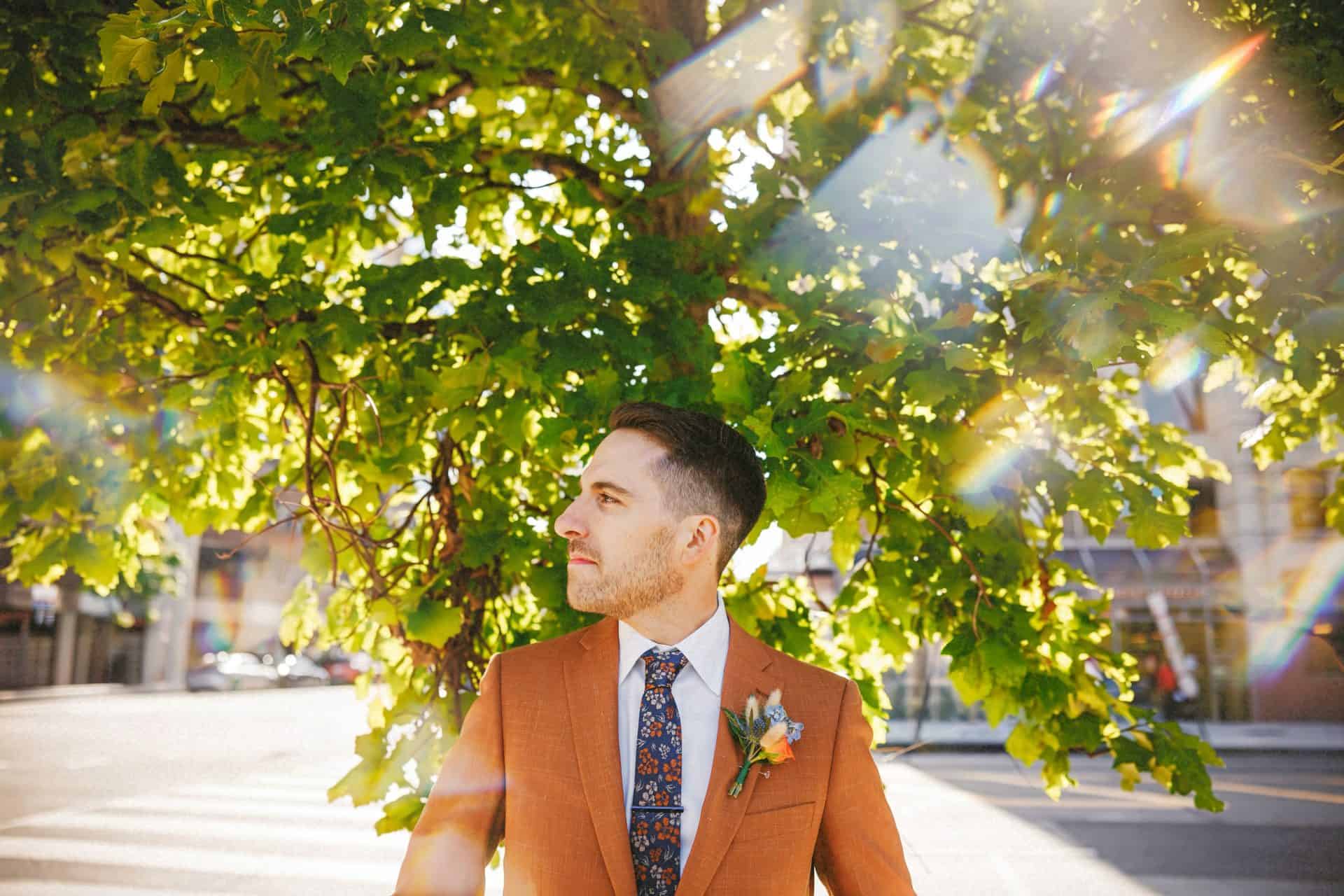 Groom in a rust suit standing under bright green leaves with rainbow lens flare on a DC street