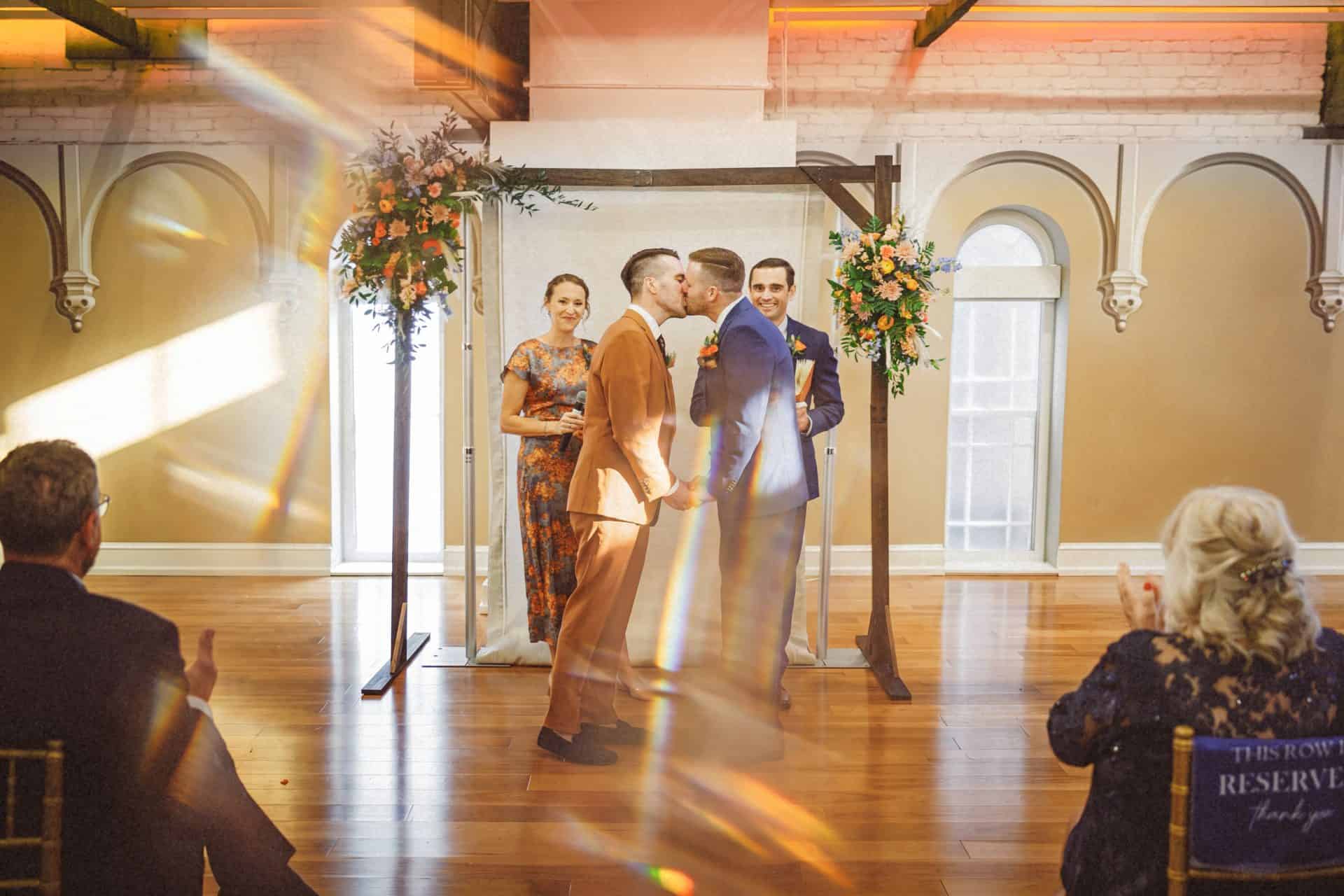 Two grooms kissing at the end of their ceremony under a floral arch at their Planet Word reception