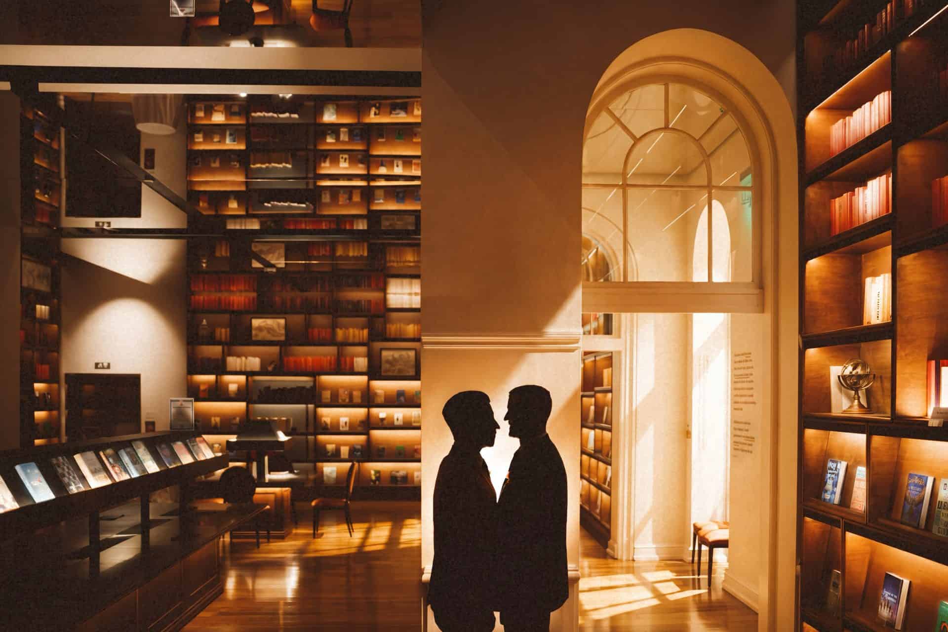 Two grooms in silhouette holding hands between glowing book walls at a Planet Word wedding in Washington DC